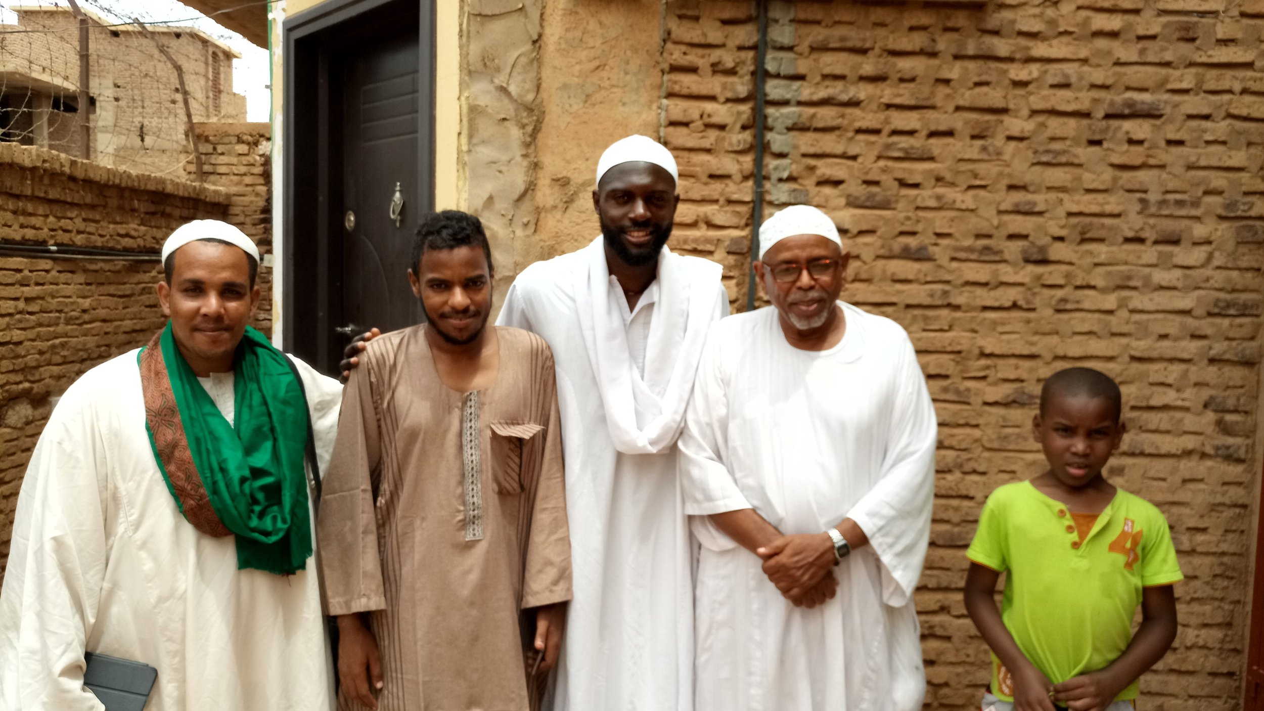 Five men and one young boy standing together outdoors in front of a brick wall, smiling at the camera. The men are dressed in traditional Islamic attire, with some wearing white robes and caps, while the boy wears a bright yellow-green shirt.