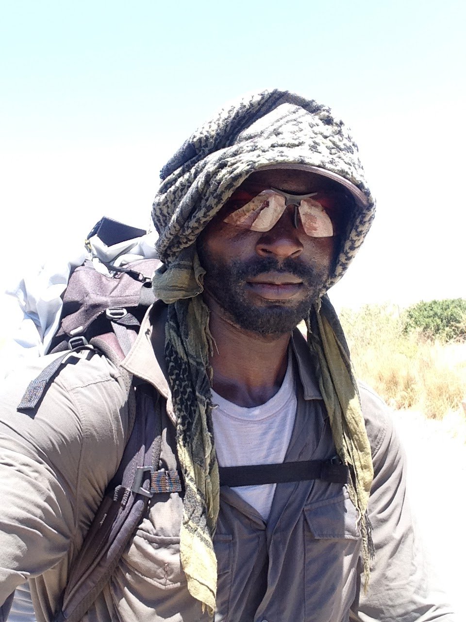 A man wearing outdoor gear, including sunglasses, a headscarf, and a backpack, standing outdoors on a sunny day.