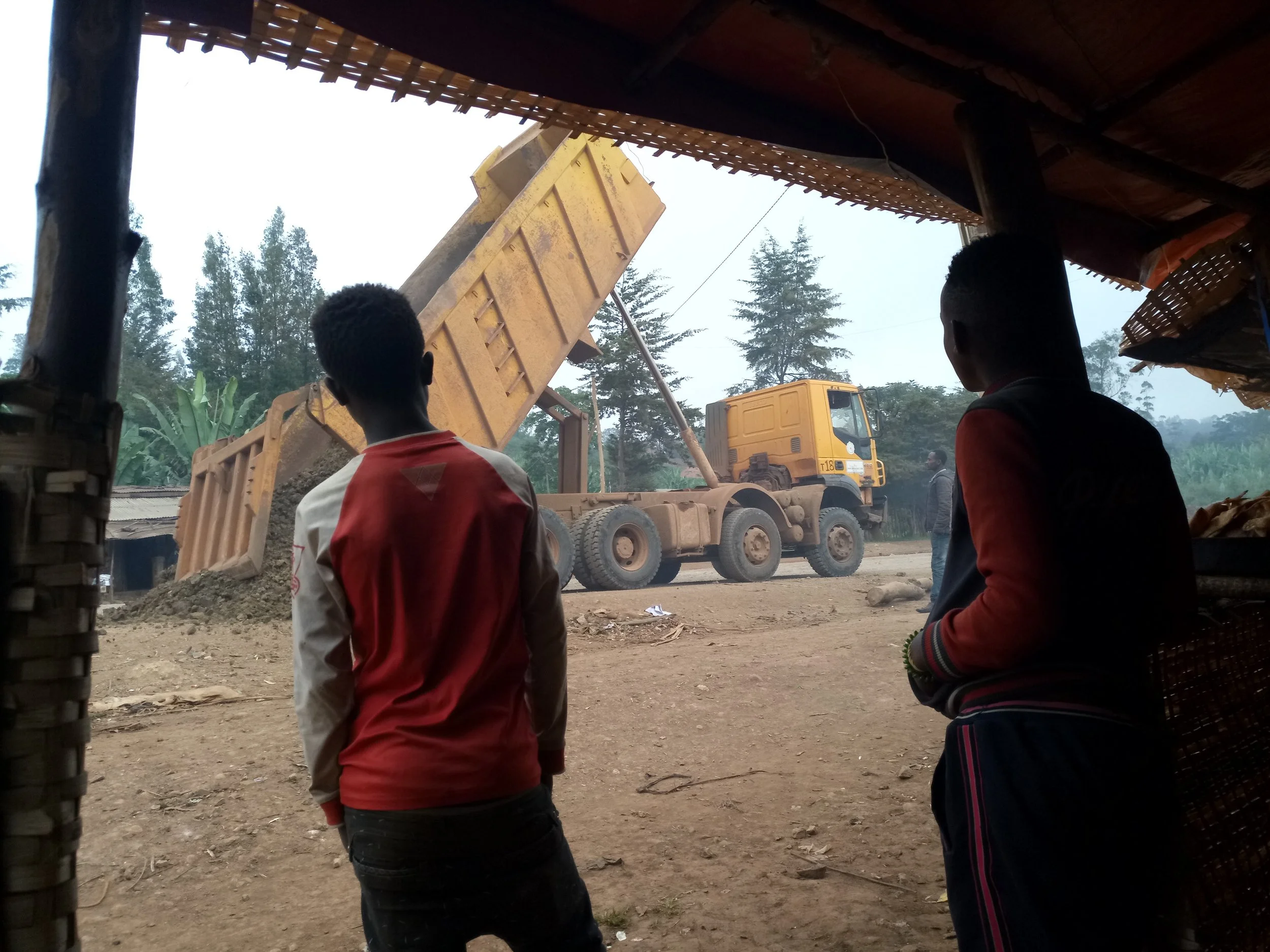 Two boys observing a yellow dump truck unloading soil or gravel in a rural area with trees in the background, viewed from inside a small shop or stall.