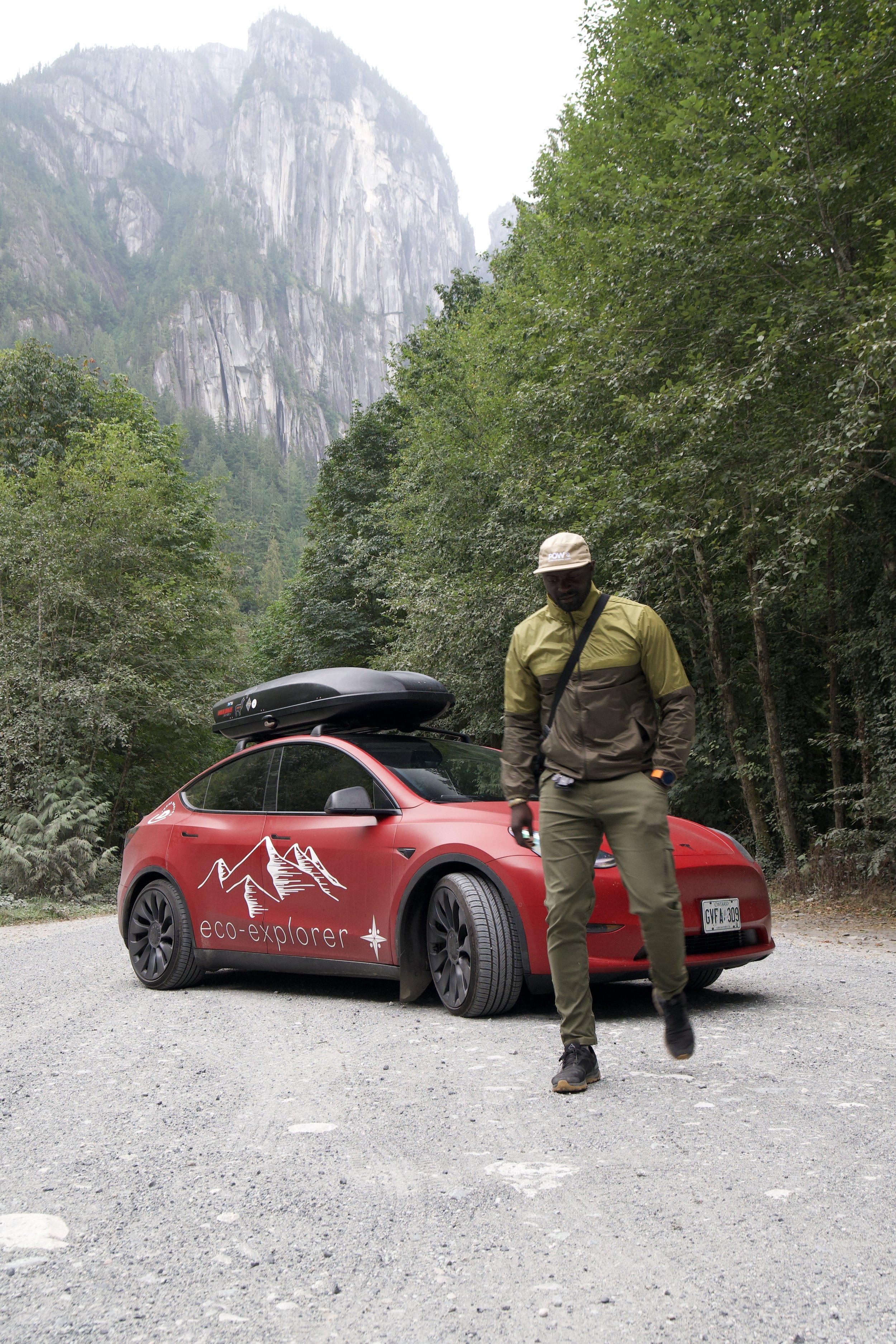 A man in outdoor gear walking on an unpaved road in a forested mountainous area, with a red electric car labeled 'eco-explorer' parked nearby.