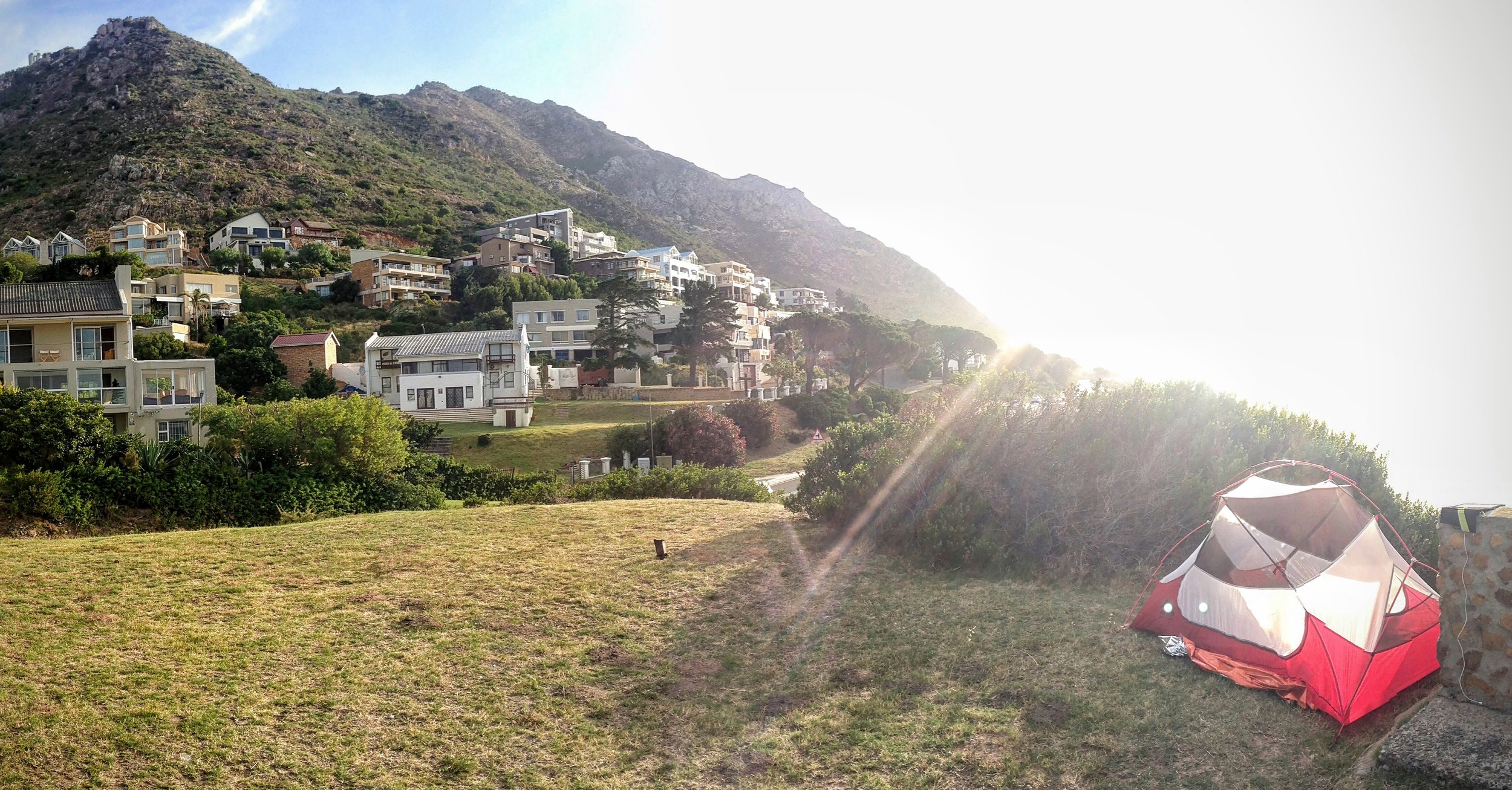 A red and white camping tent on a grassy hillside with residential houses on a mountain slope in the background, and sunlight streaming over the scene.