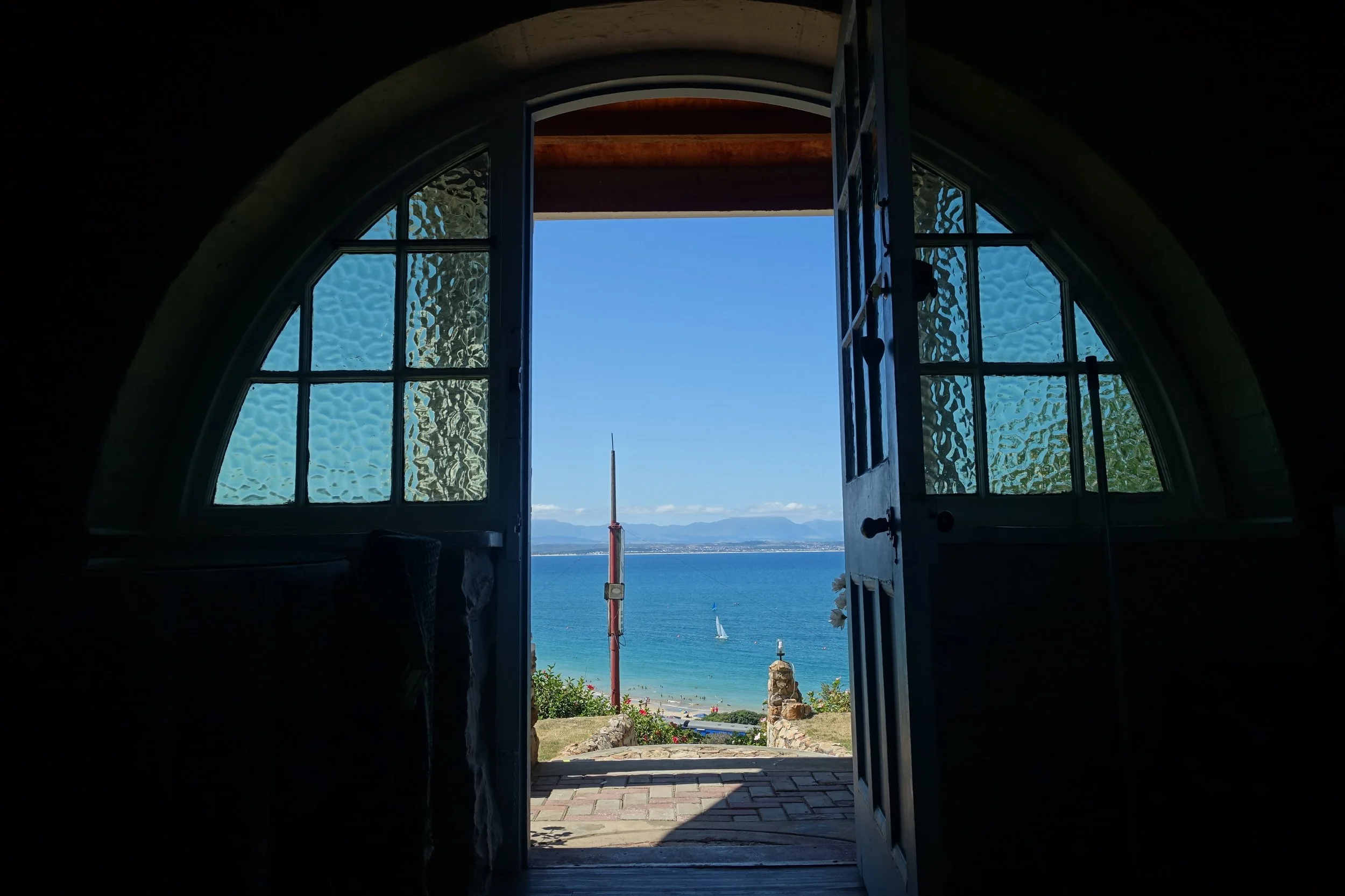 View through a church doorway showing a beach, ocean, sailboat, and distant mountains under a blue sky.