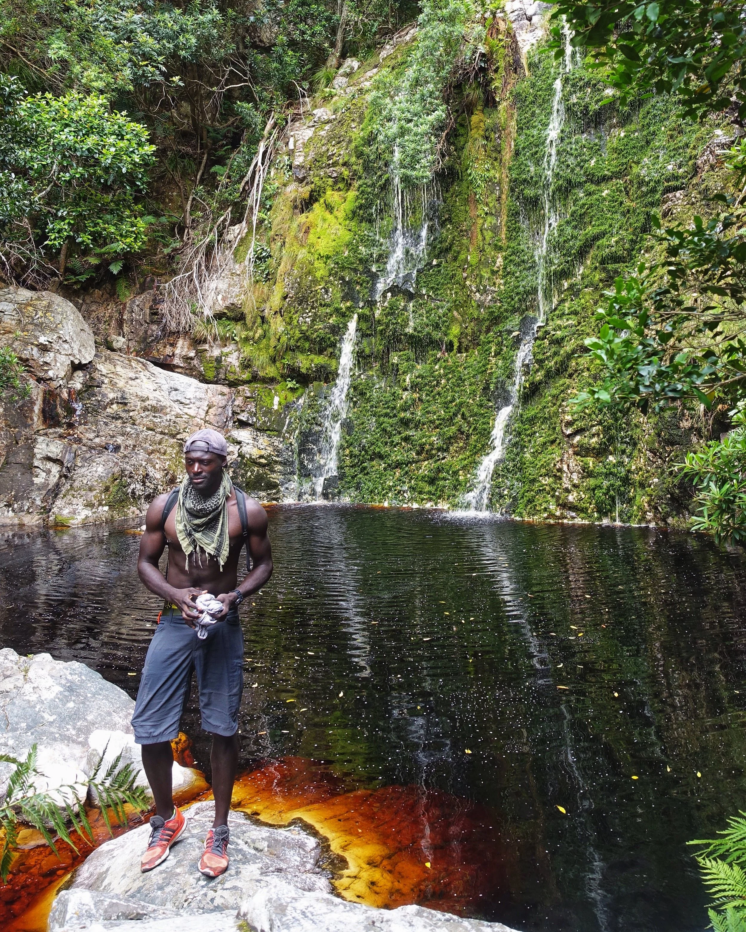 A shirtless man wearing a gray cap, scarf, and gray shorts stands on a rock by a dark pool of water near a moss-covered waterfall in a lush green forest.