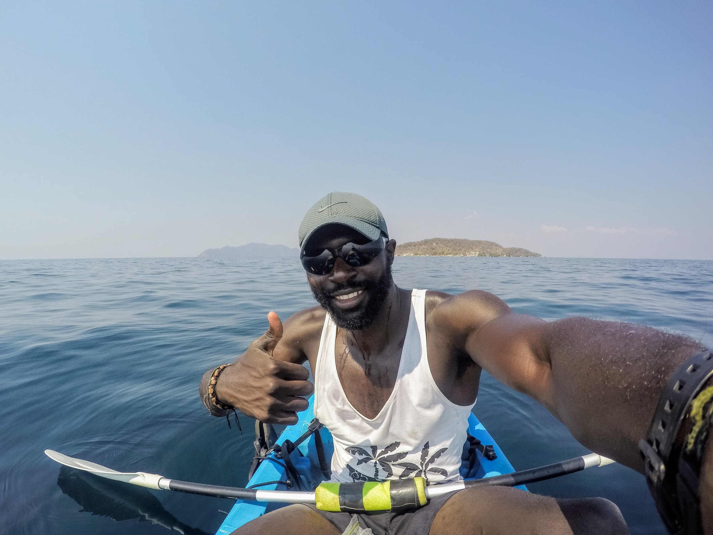 Man kayaking on the ocean wearing sunglasses, a cap, tank top, and wristbands, giving a thumbs up while smiling.