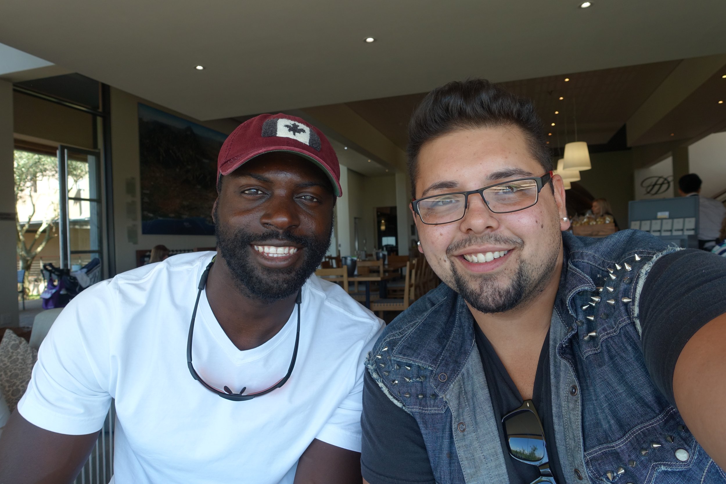Two men smiling for a selfie in a restaurant, one wearing a white t-shirt and red cap, and the other wearing glasses and a denim vest.