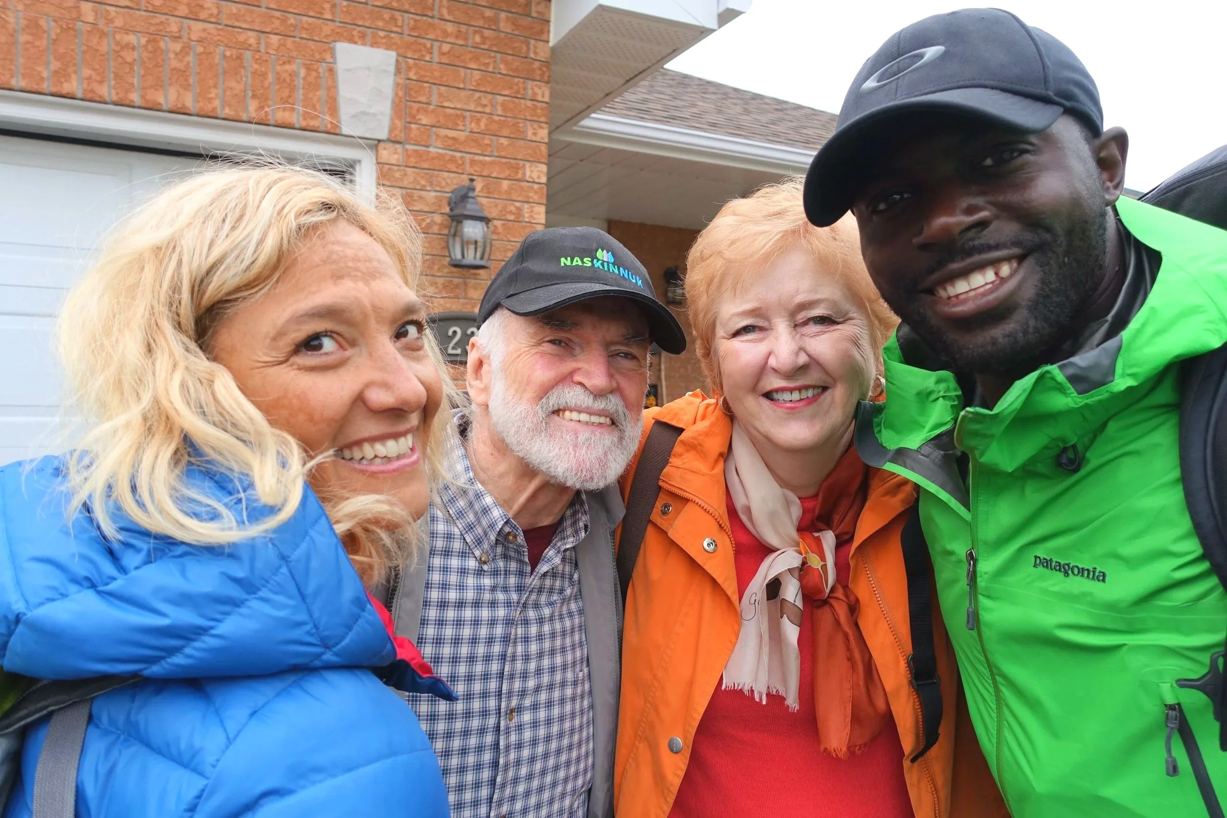 Four people smiling outdoors in front of a brick house, dressed in outdoor jackets and hats.