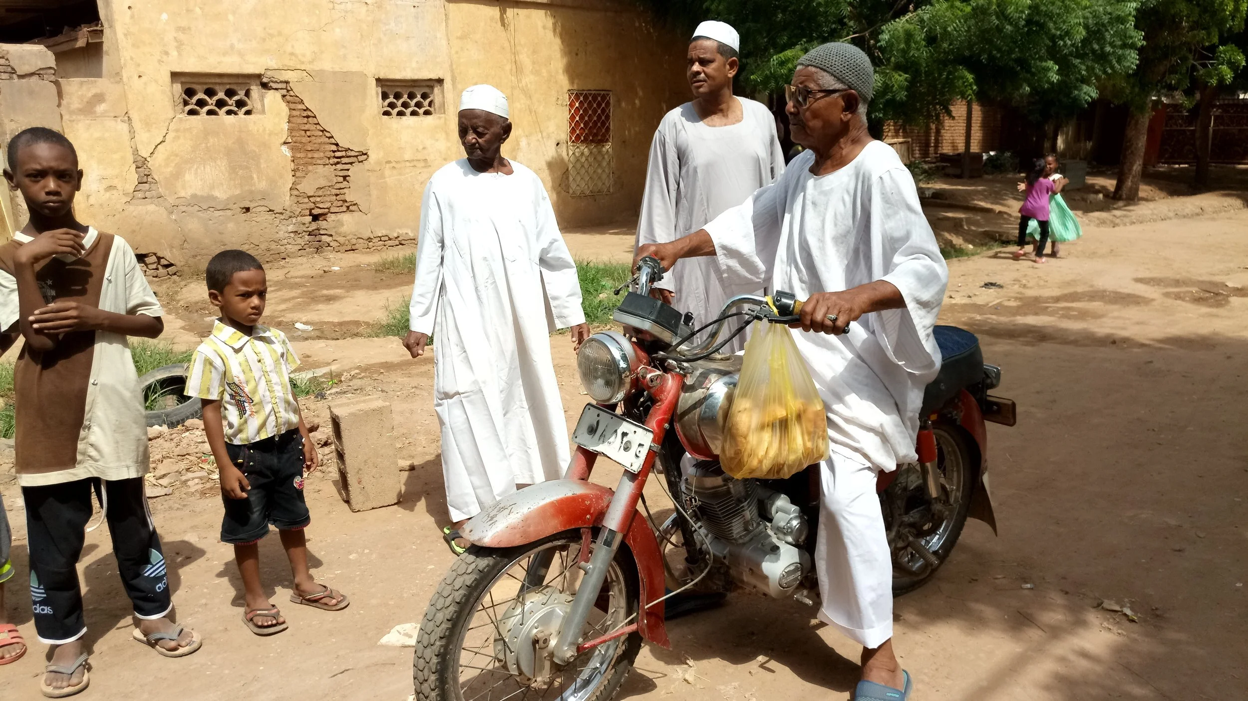 Group of men and children standing outdoors, with an elderly man sitting on a motorcycle holding a plastic bag with what appears to be food, while three children and two other men look on. The scene shows an old building with a weathered wall in the 
