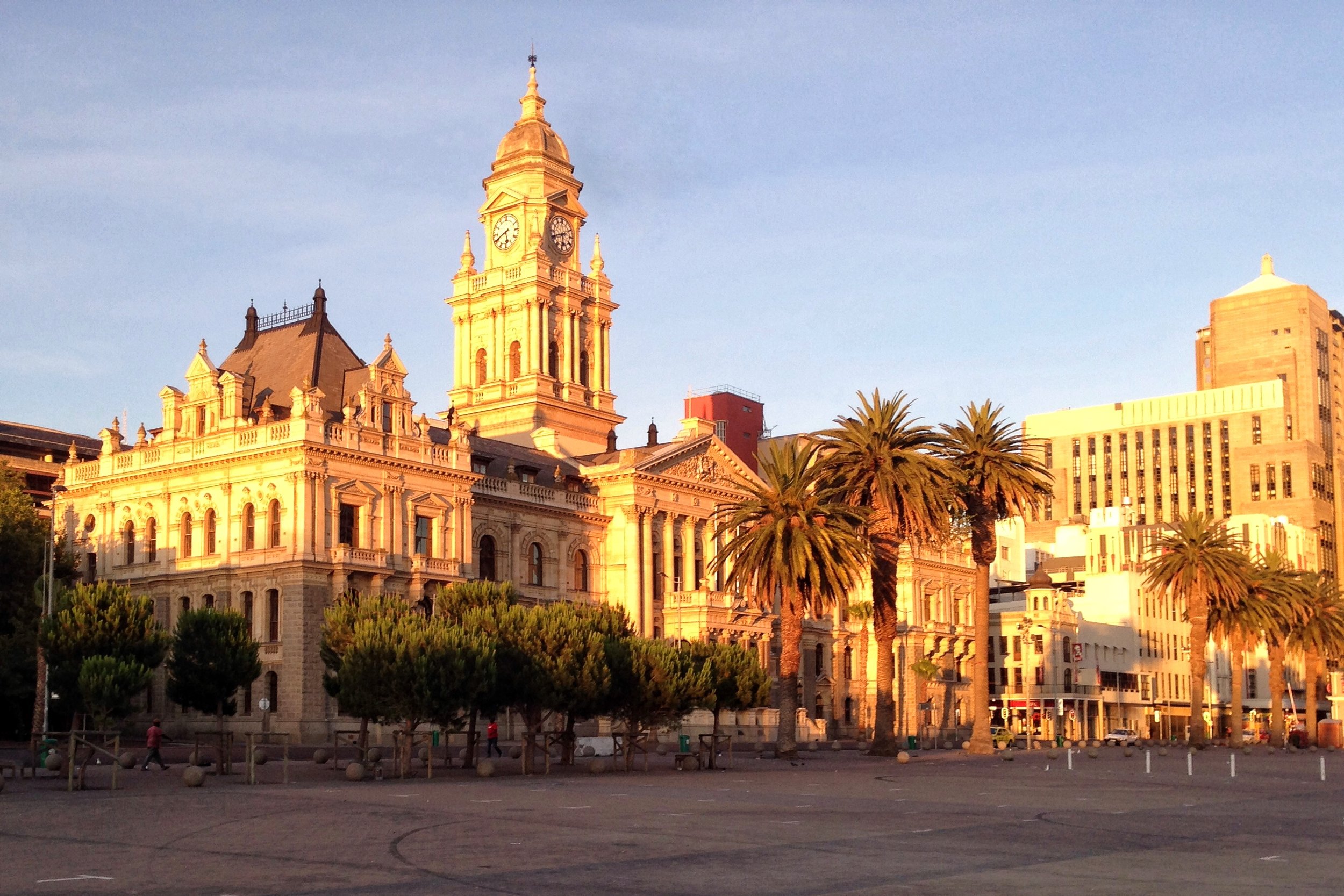 A historic building with a clock tower and ornate architecture, surrounded by palm trees and modern buildings, bathed in warm sunlight.