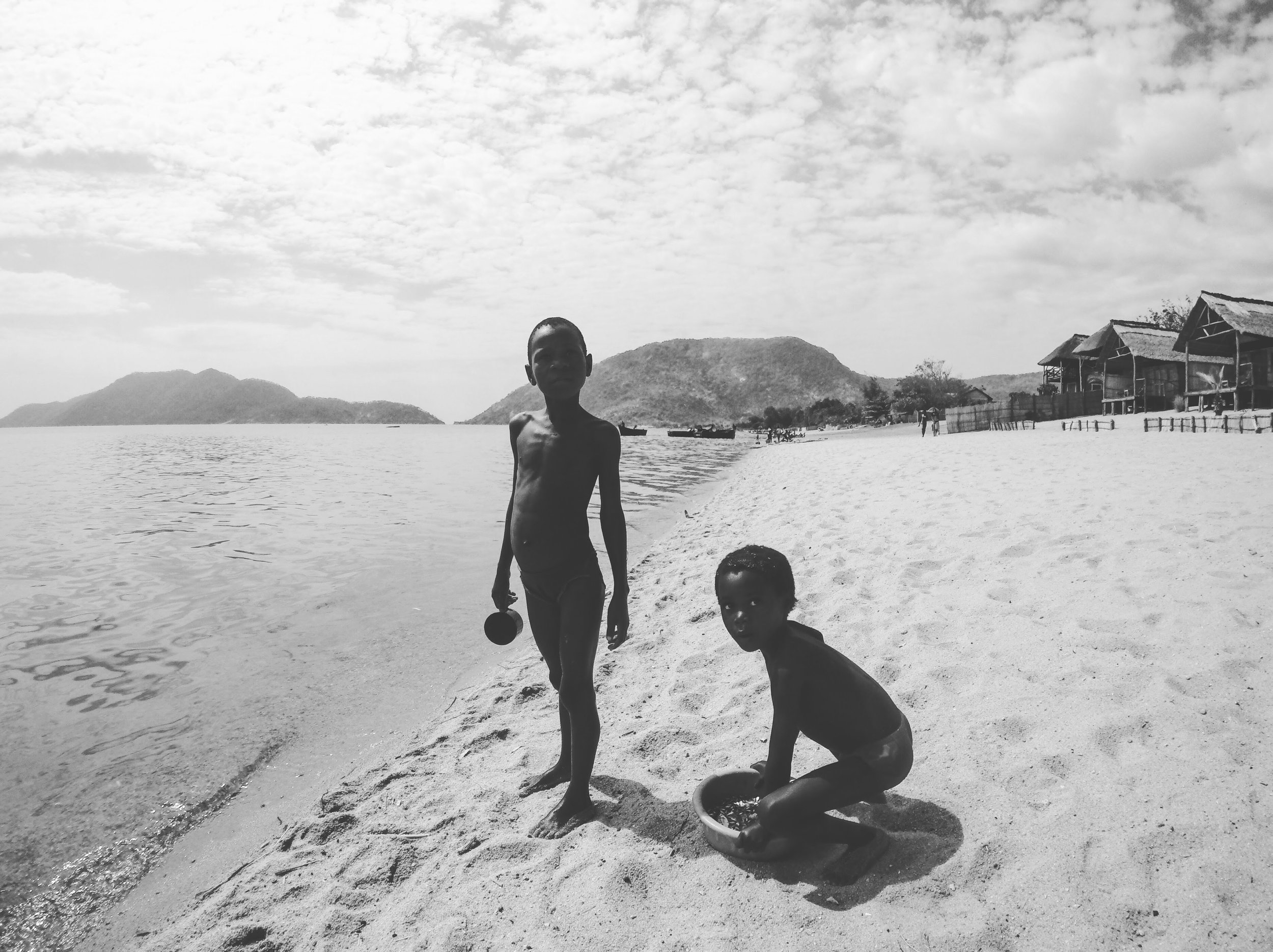 Two children playing on a sandy beach near water, with mountains and houses in the background, black and white photograph.
