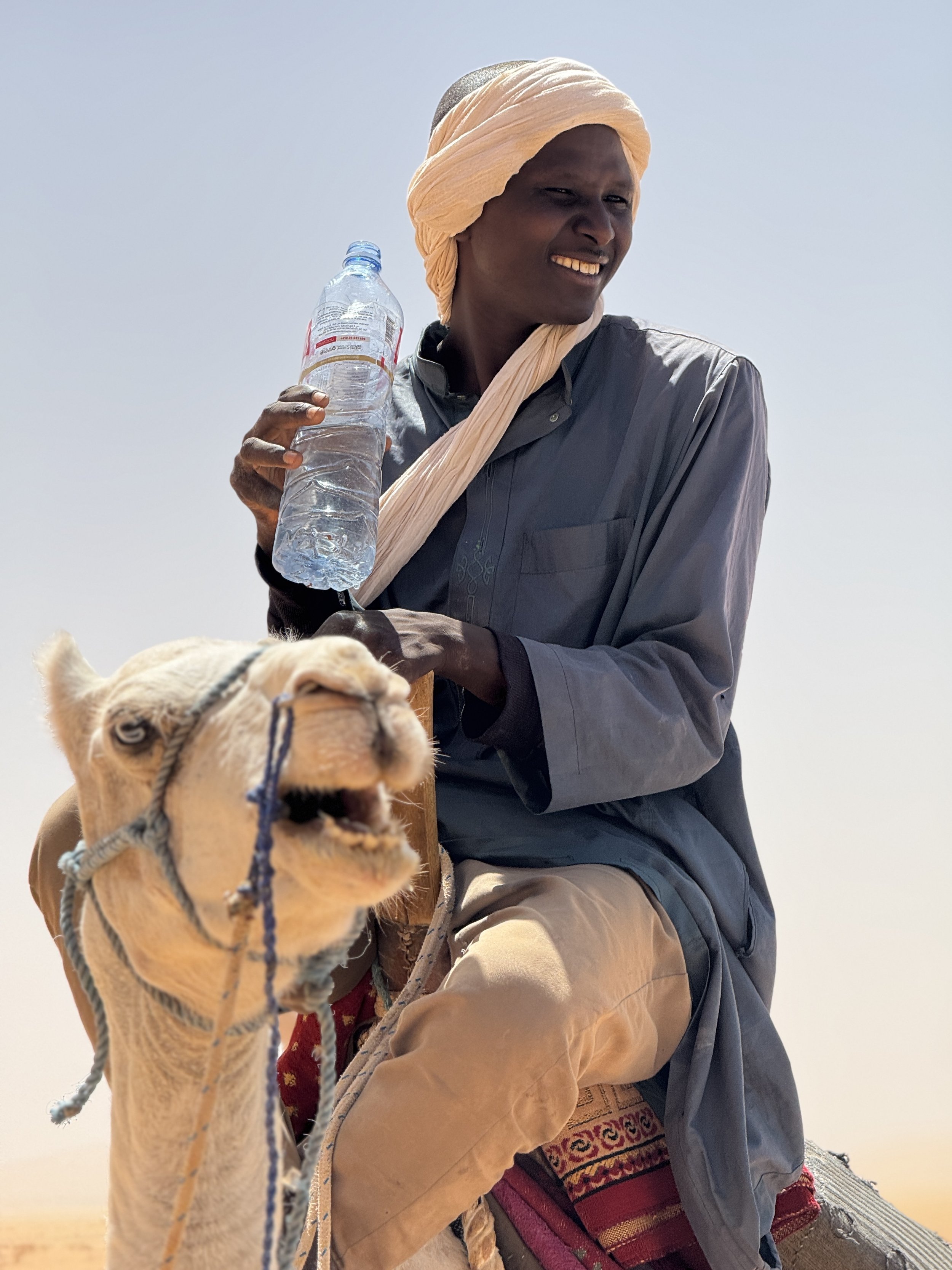 A man riding a camel in a desert, holding a bottle of water, wearing a headscarf, with a clear sky in the background.