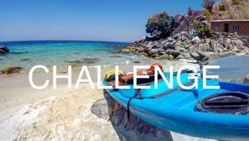 A blue kayak on a sandy beach with clear ocean water and rocks in the background.