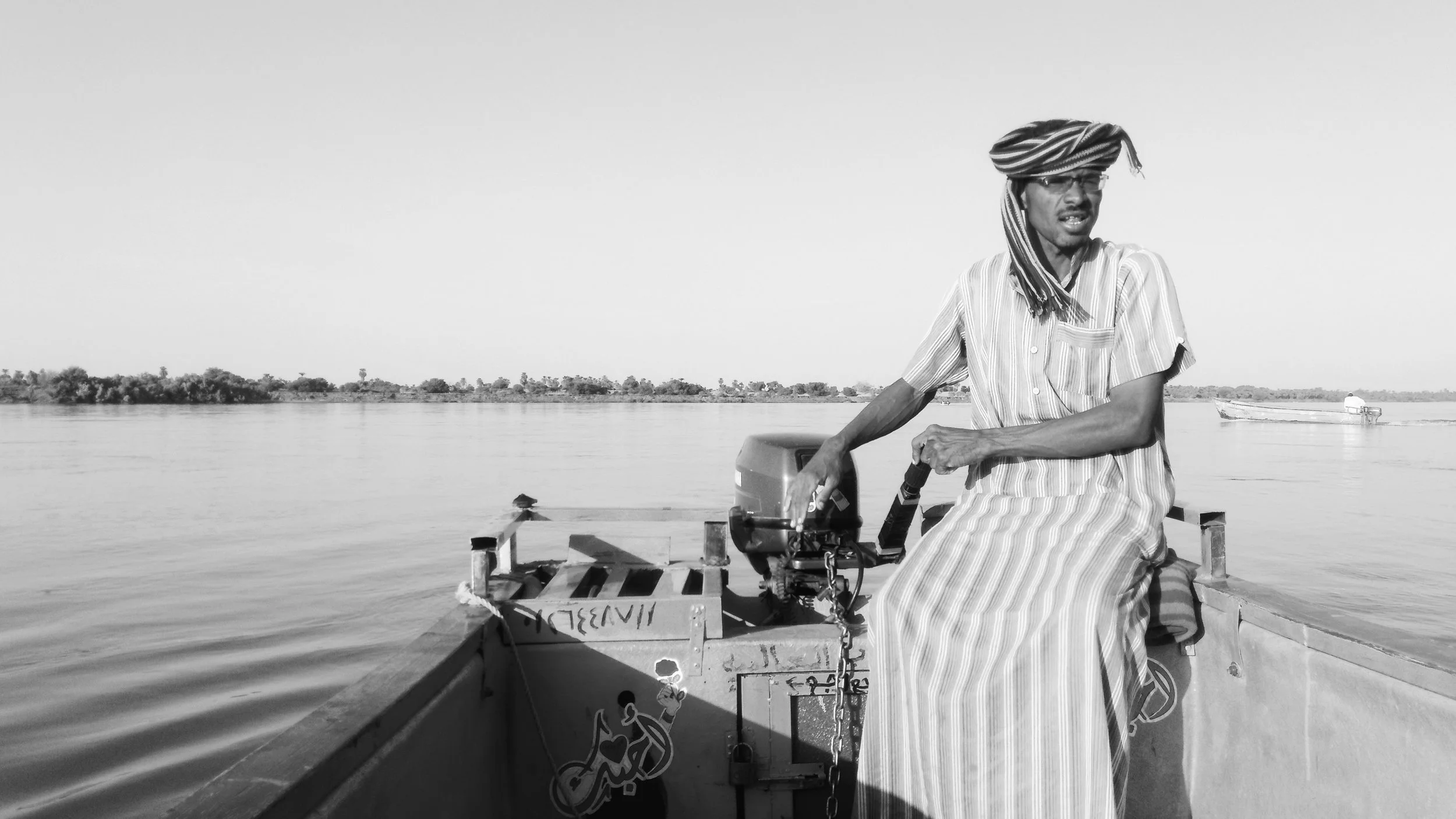 A man wearing a striped head wrap and a striped traditional garment sitting in a boat on a river, operating an outboard motor, with a distant boat and land visible in the background.