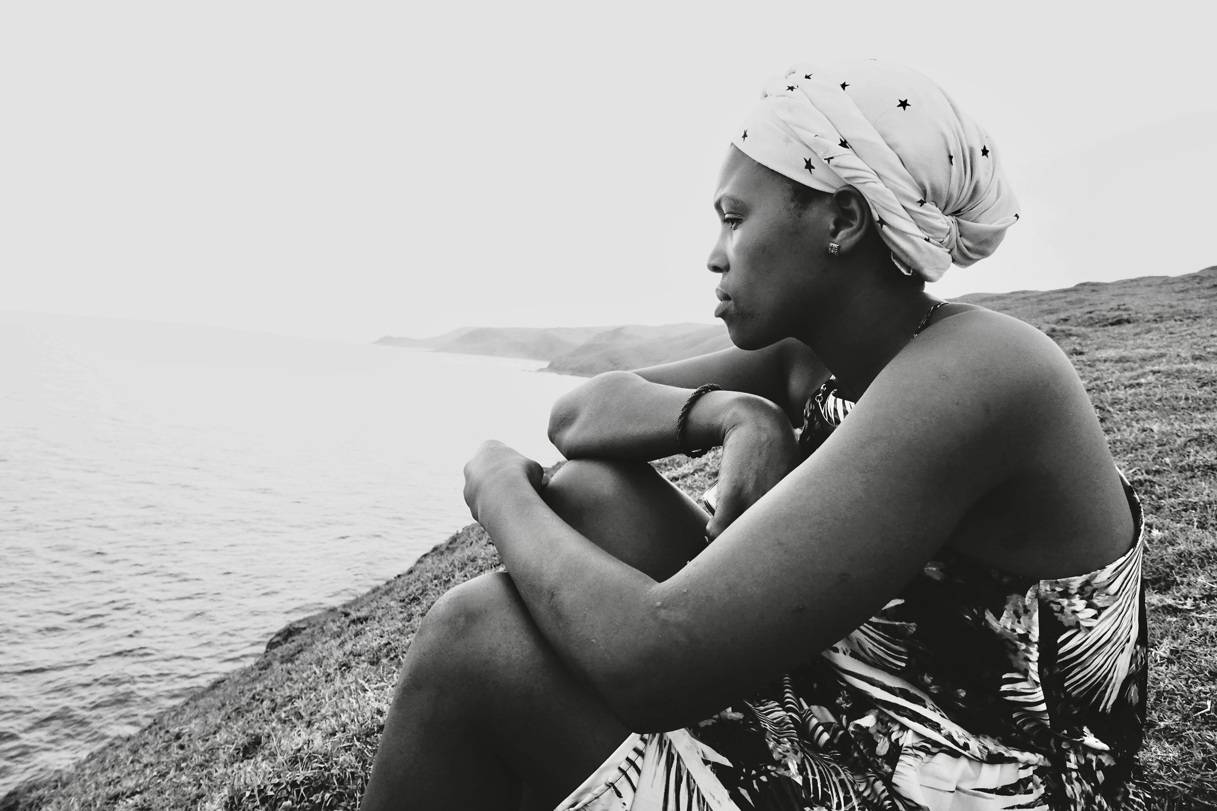 A woman sitting on a grassy hill near the water, looking contemplative, wearing a patterned dress and a headscarf.