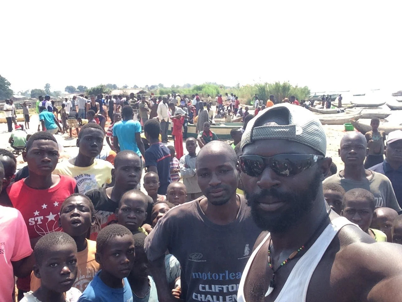 A large group of children and men gathered on a sandy shoreline near boats, with some boats on water and others on land, under a bright sky during daytime.