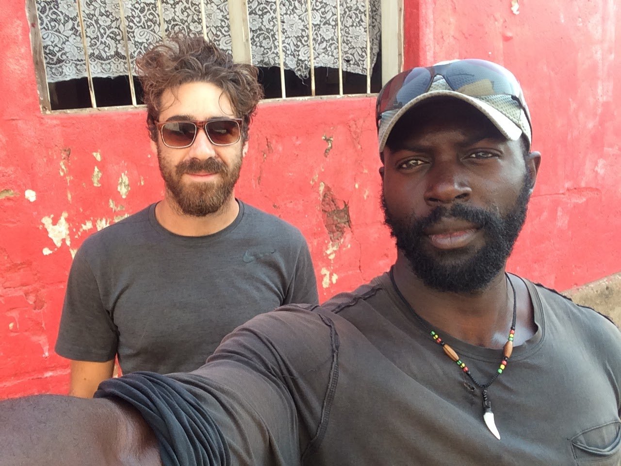Two men posing for a selfie in front of a worn red wall with a barred window. The man in the foreground wears a gray cap, sunglasses on his head, a gray shirt, and a beaded necklace with a shell pendant. The man in the background has curly hair, a be