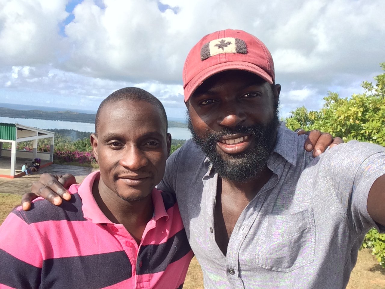 Two men posing outdoors with a scenic landscape of water, trees, and cloudy sky in the background. One man is wearing a pink and navy striped polo shirt, the other is wearing a light gray shirt and a red cap. They are smiling with one man's arm aroun