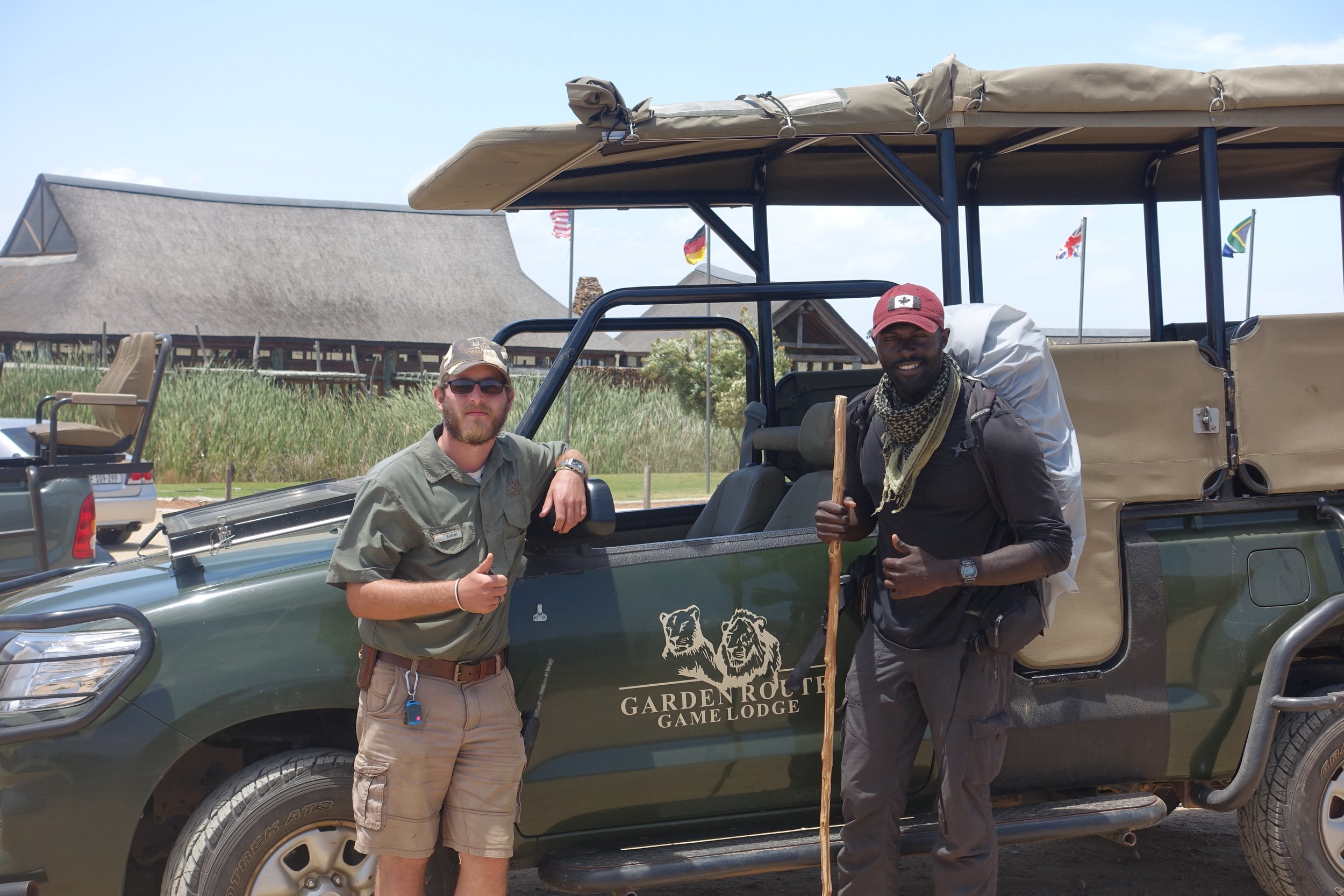 Two men standing beside a green game lodge vehicle at GARDENROCK GAME LODGE, one giving a thumbs up and the other holding a wooden stick, with flags and a building in the background.