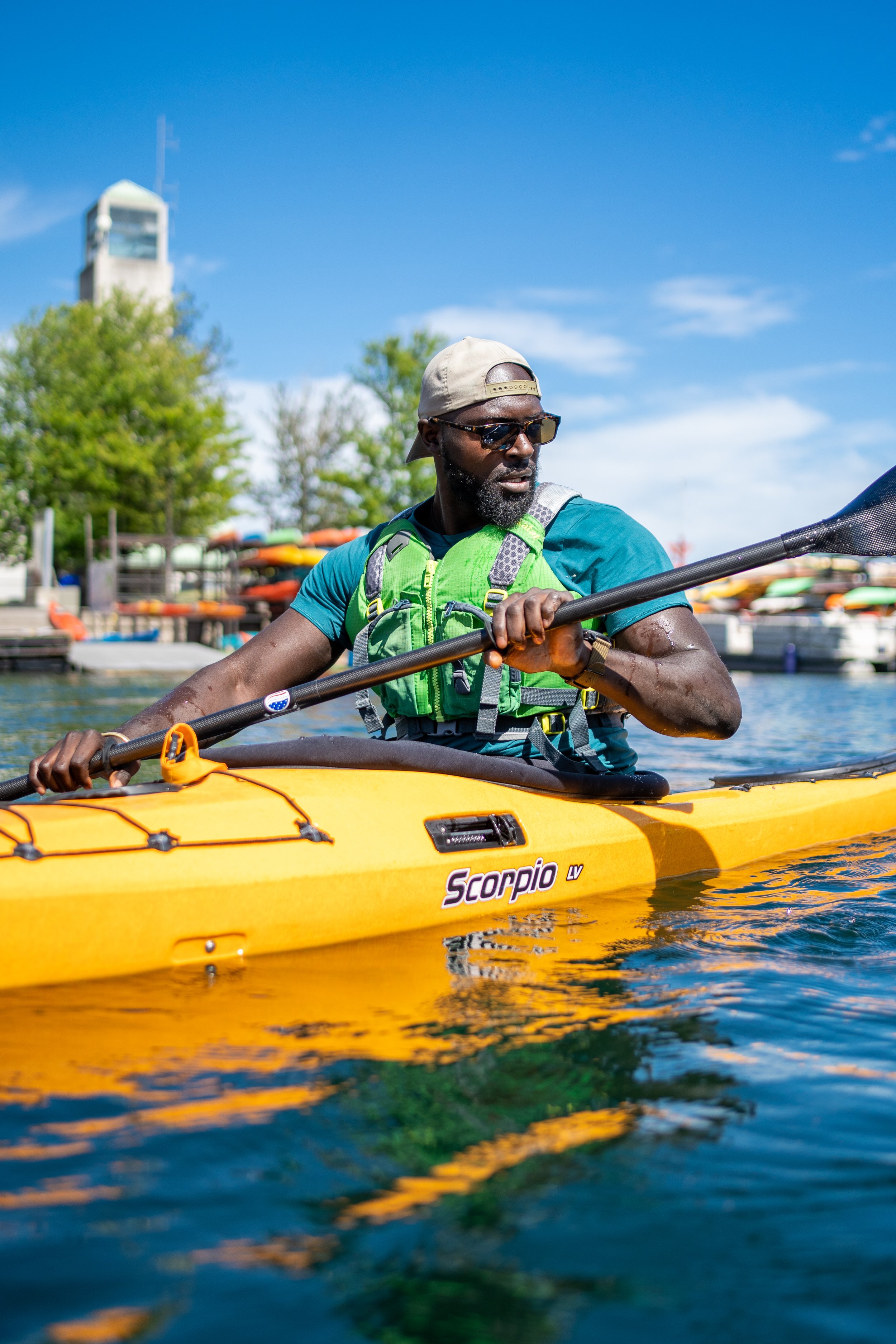 A man kayaking on a body of water, wearing sunglasses, a beige cap backwards, a blue shirt, and a green life jacket. There are boats and kayaks in the background, and the sky is clear and blue.