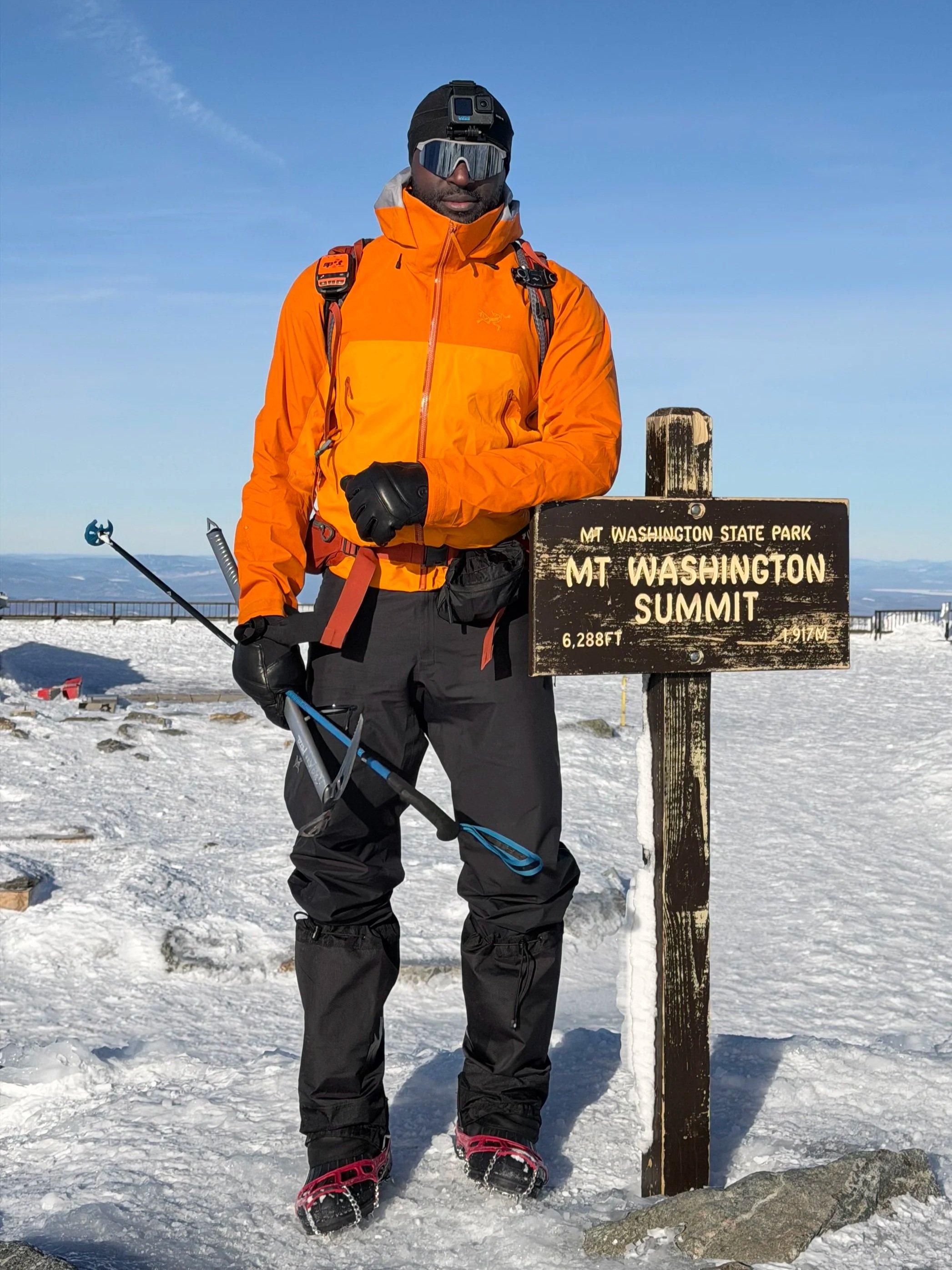 A person standing on snow-covered ground at Mount Washington Summit, dressed in orange and black winter gear, holding a hiking pole, next to a sign that reads 'Mt Washington State Park, Mt Washington Summit, 6,288 FT 1,917 M'.