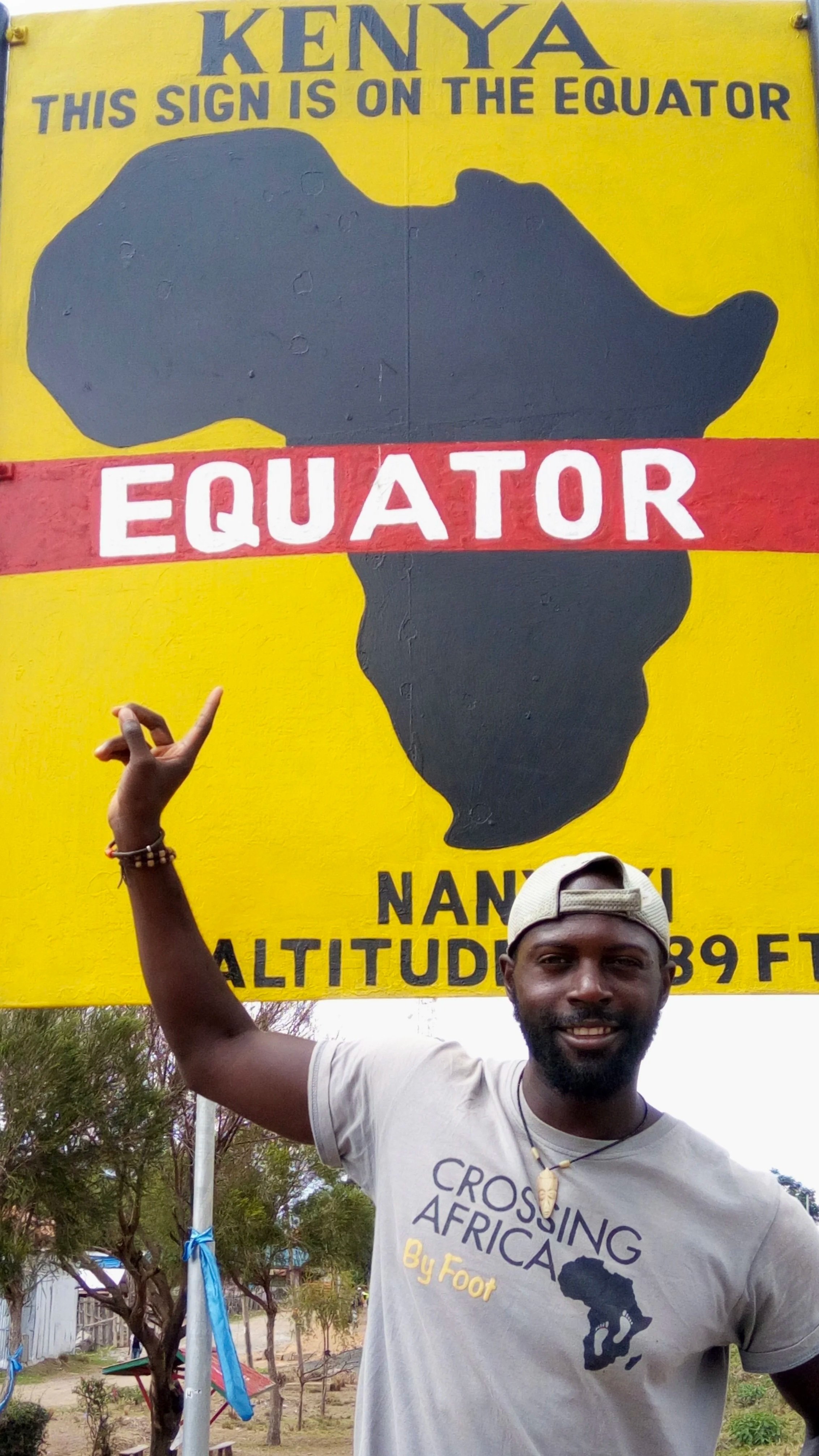 Man standing in front of a yellow sign that says, "Kenya. This sign is on the equator," with a black silhouette of Africa, and the word "Equator" written across it. The man is smiling, pointing at the sign with his right hand, wearing a light gray t-shirt that says "Crossing Africa by Foot" with a map of Africa, a white cap, and a seashell necklace.