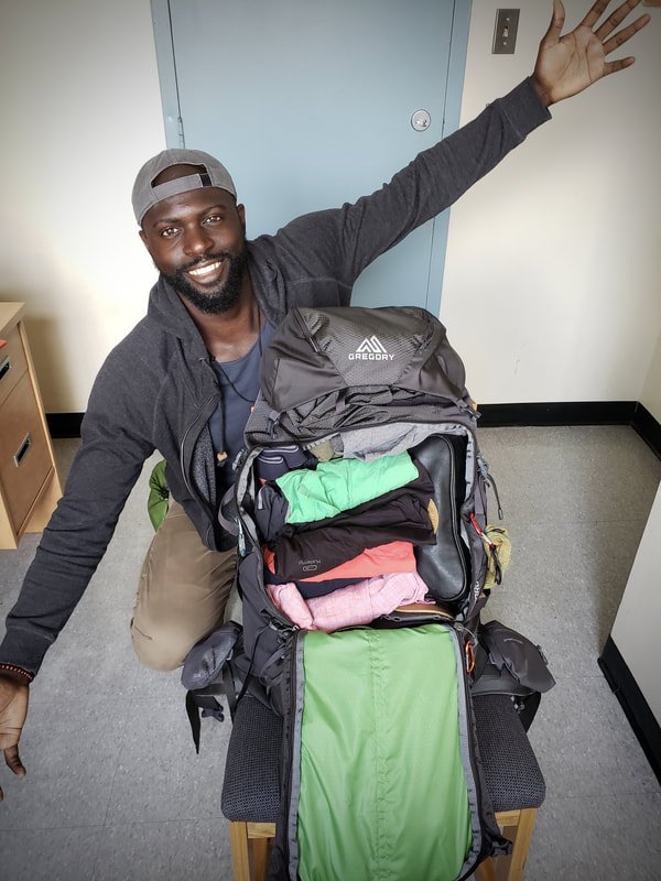 A smiling man wearing a cap and gray jacket kneeling beside an open hiking backpack filled with clothing and gear.