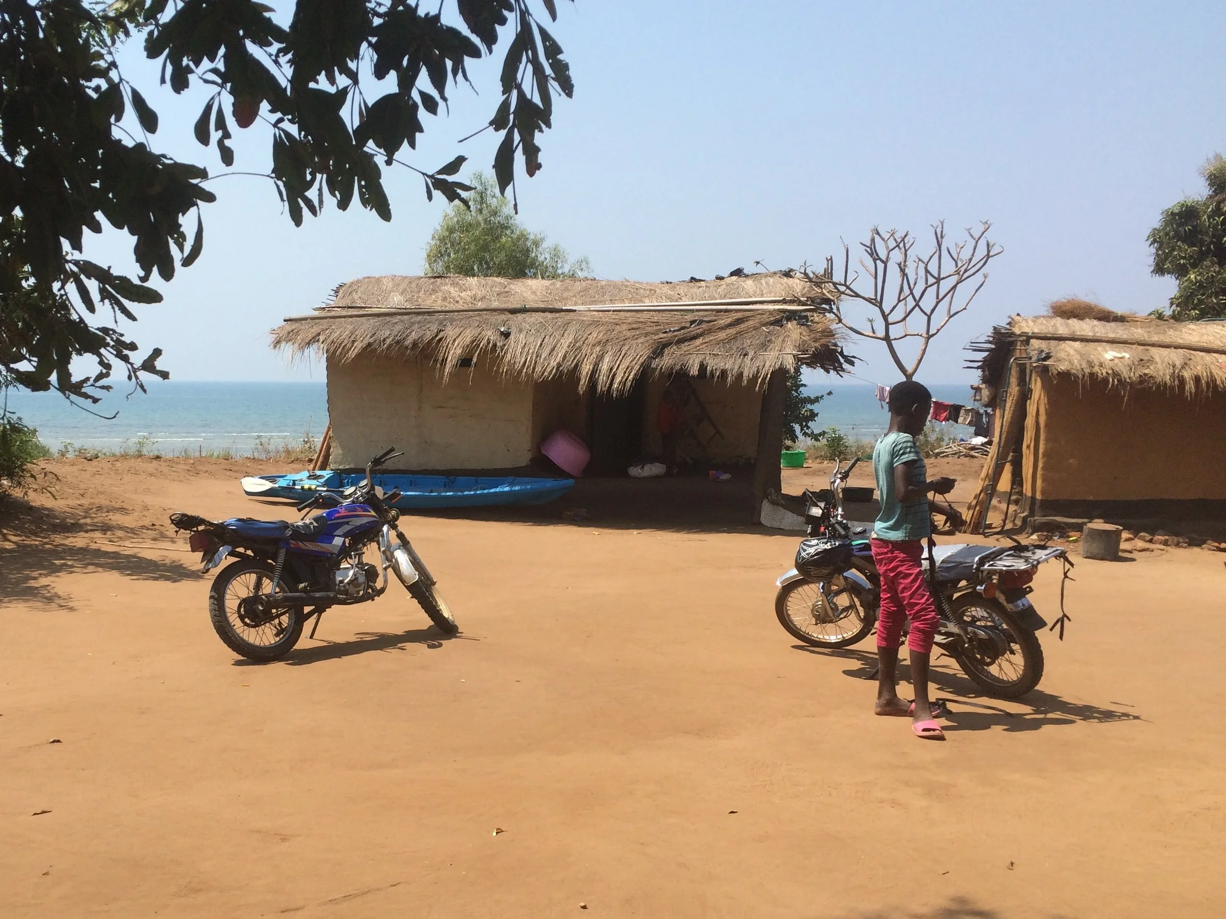 A young person standing next to a motorcycle on a dirt road near a beach, with huts made of thatched roofs and a boat in the background.