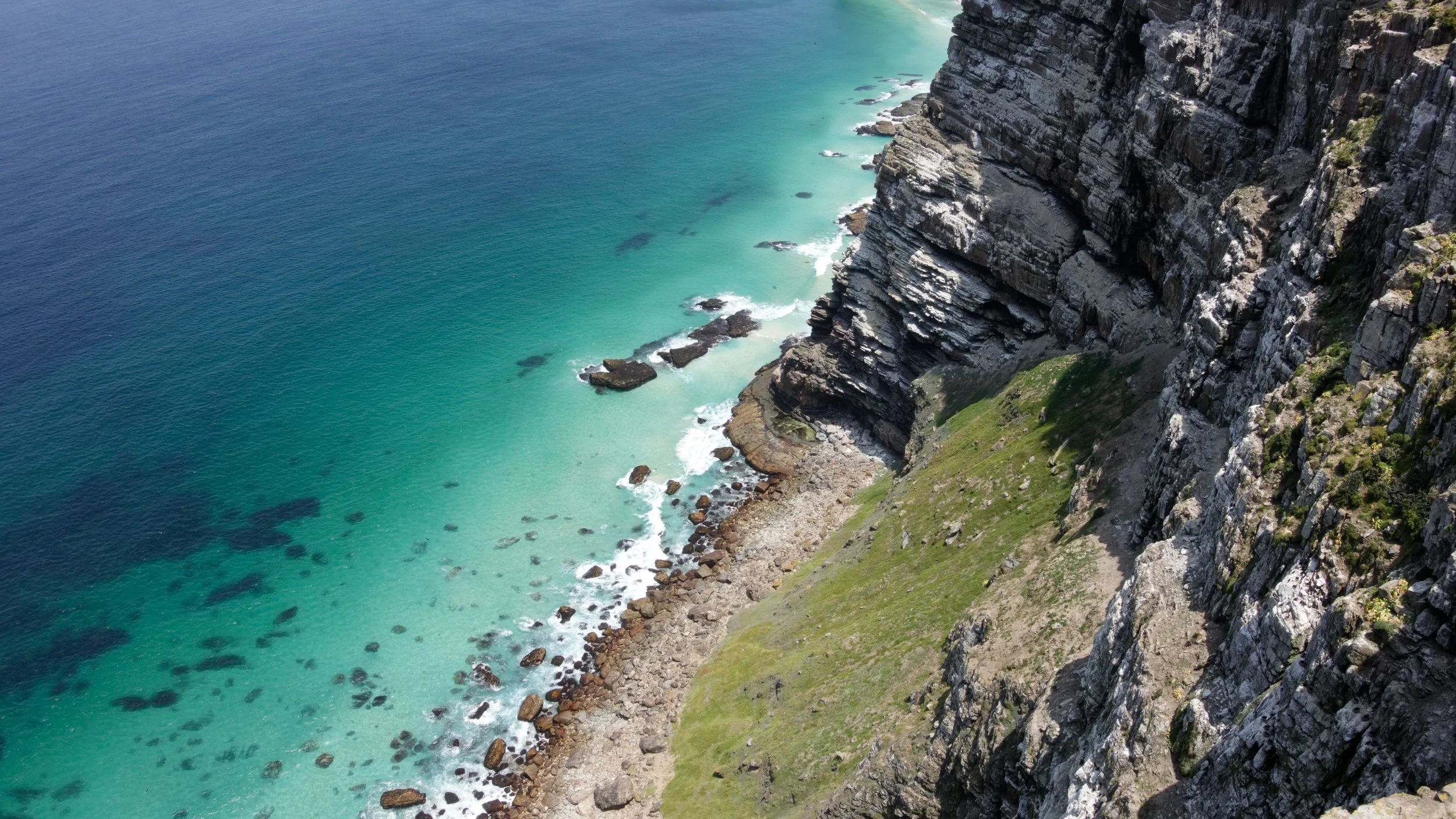 Aerial view of a rugged coastline with steep cliffs, grassy patches, and turquoise ocean waters.
