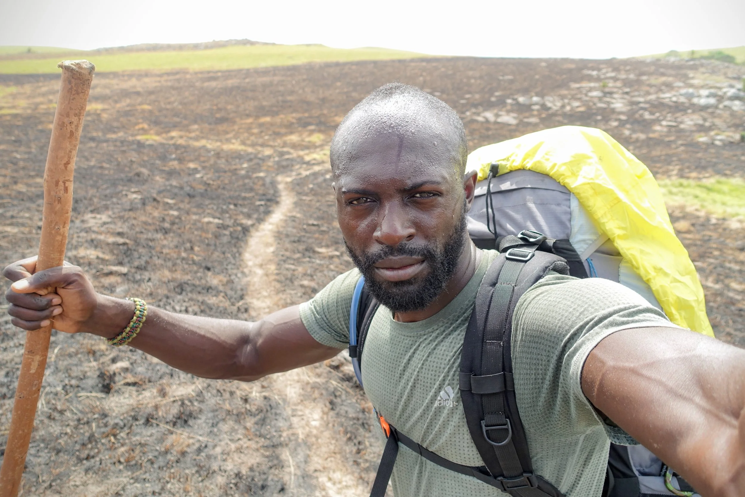 Man taking a selfie with a hiking trail and field in the background, wearing a backpack and holding a wooden stick.