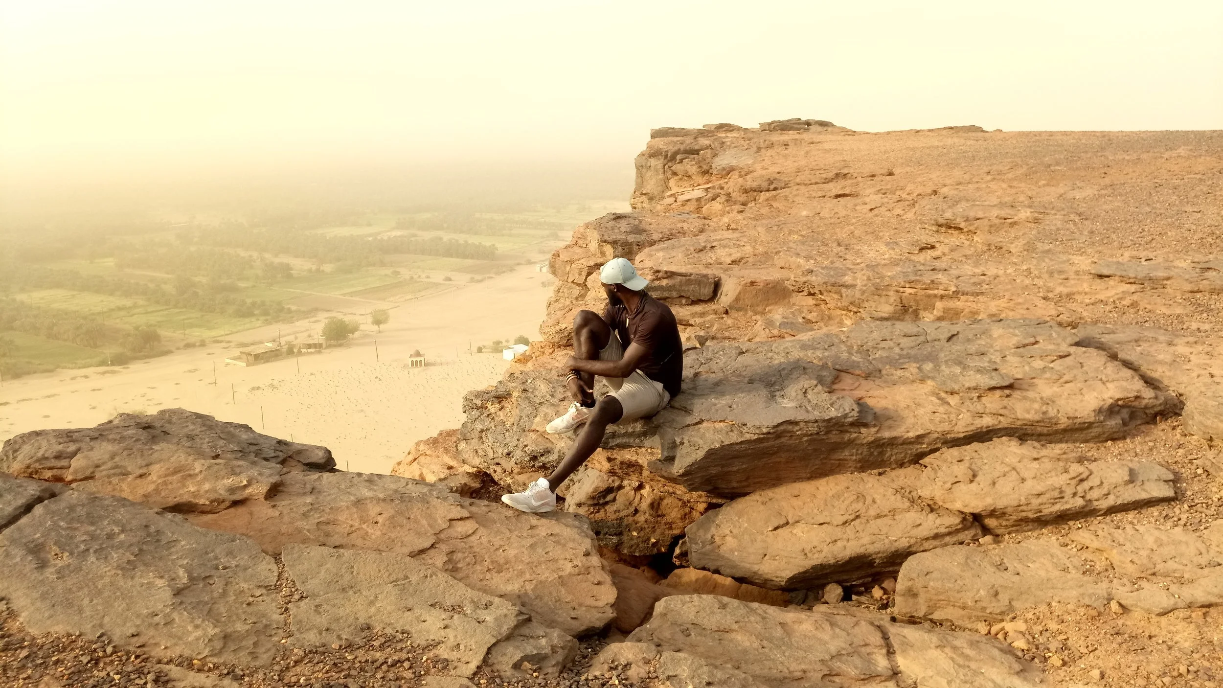 A man wearing a white cap, black t-shirt, beige shorts, and white sneakers sitting on rocks at the edge of a cliff with a desert landscape below on a hazy day.