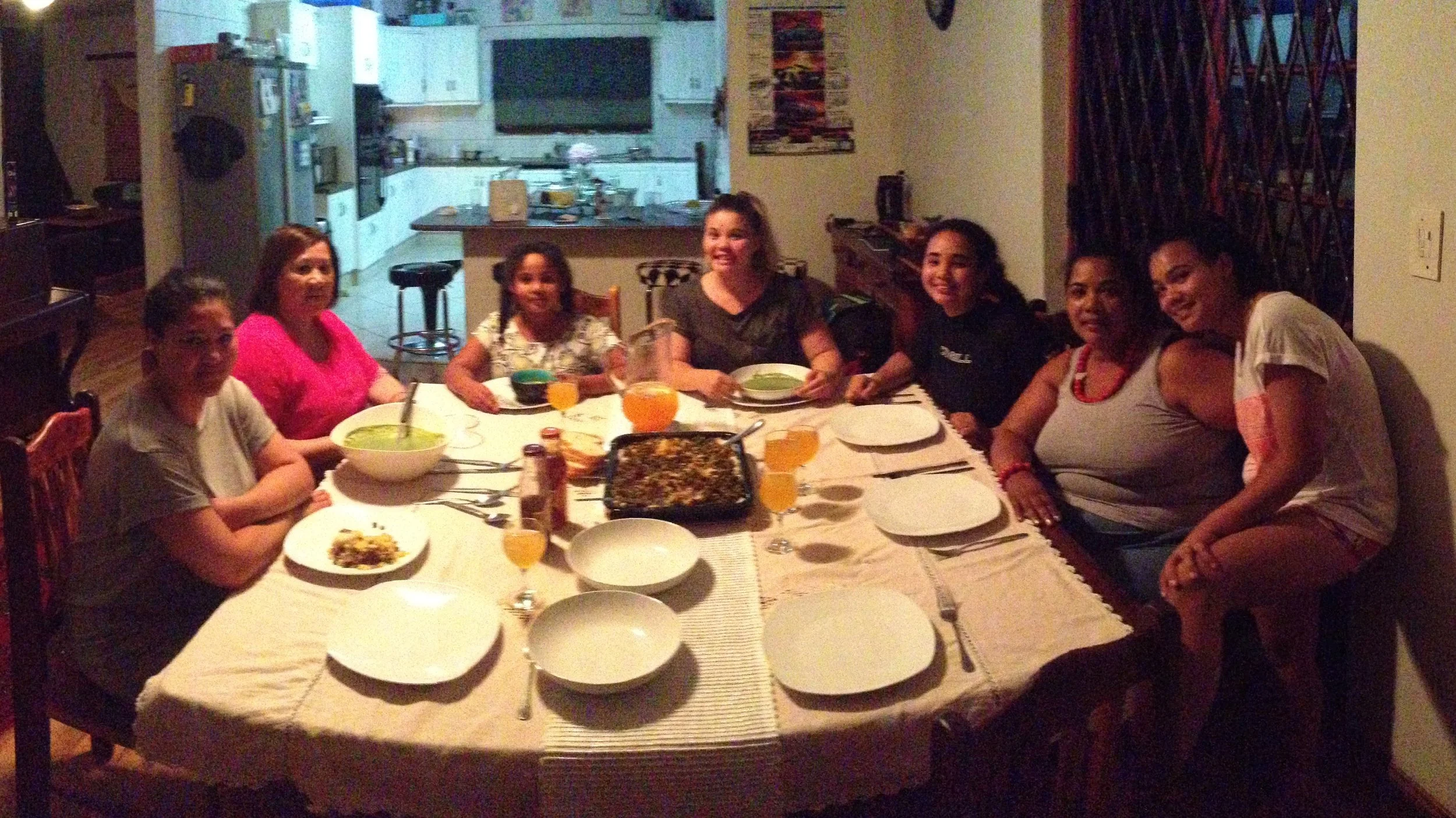 A group of nine people, mostly women and one girl, gathered around a dining table with plates, glasses, and food in a warmly lit home dining room.