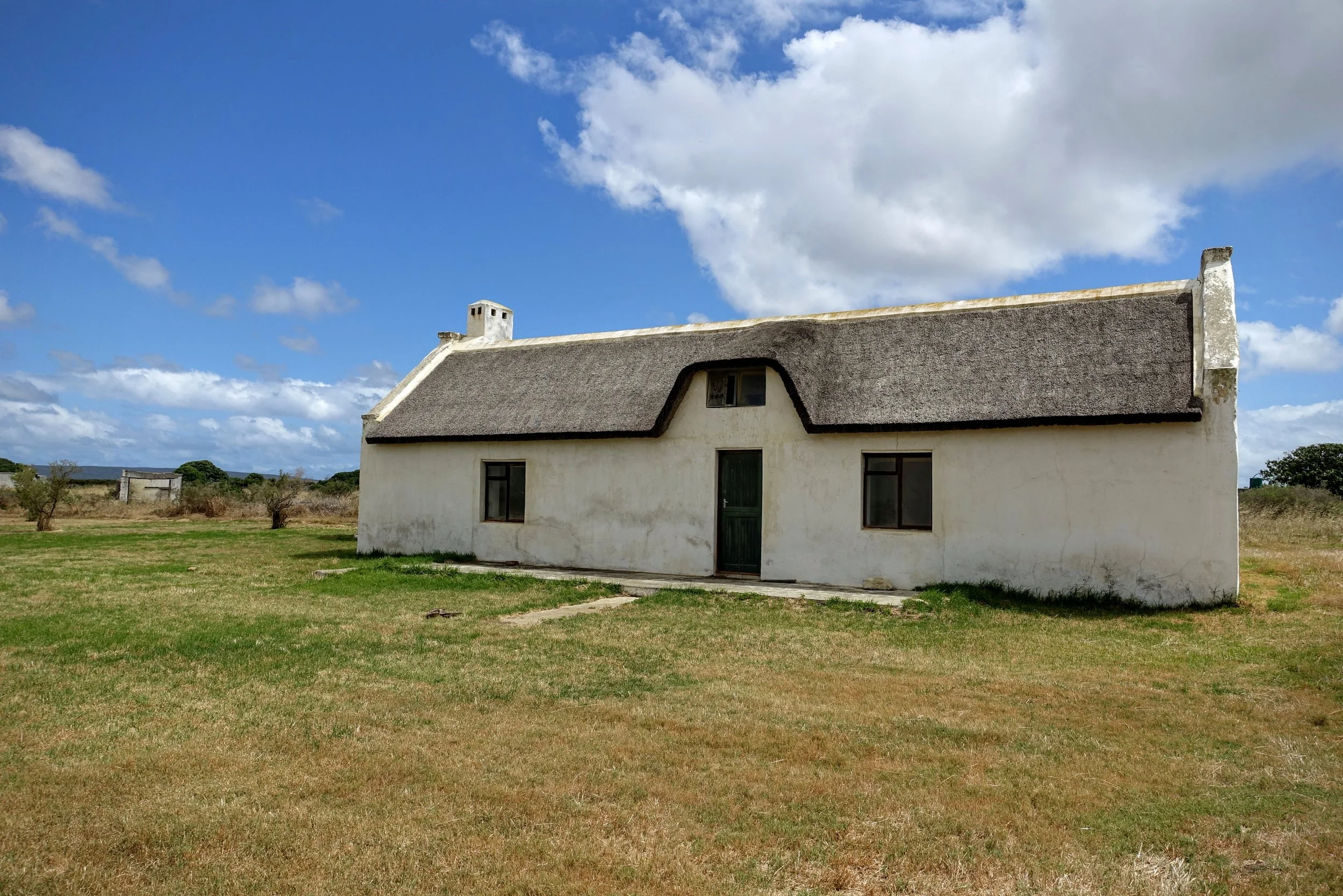A white house with a thatched roof in an open grassy field under a partly cloudy sky.