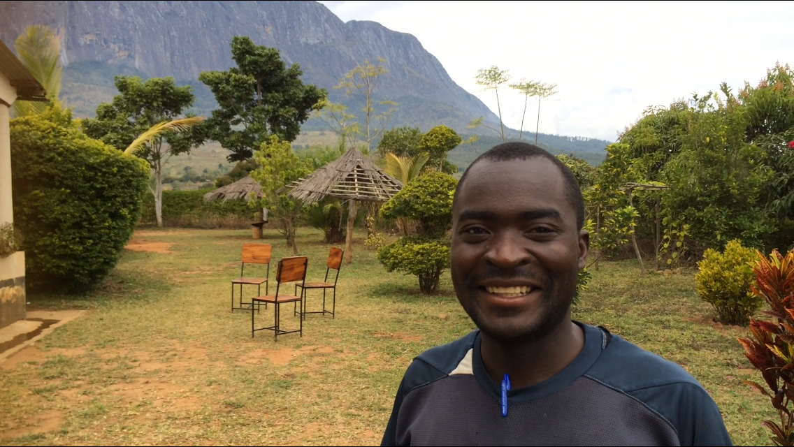 A smiling man outdoors in a lush garden with trees, bushes, and thatched umbrellas, with mountains in the background.