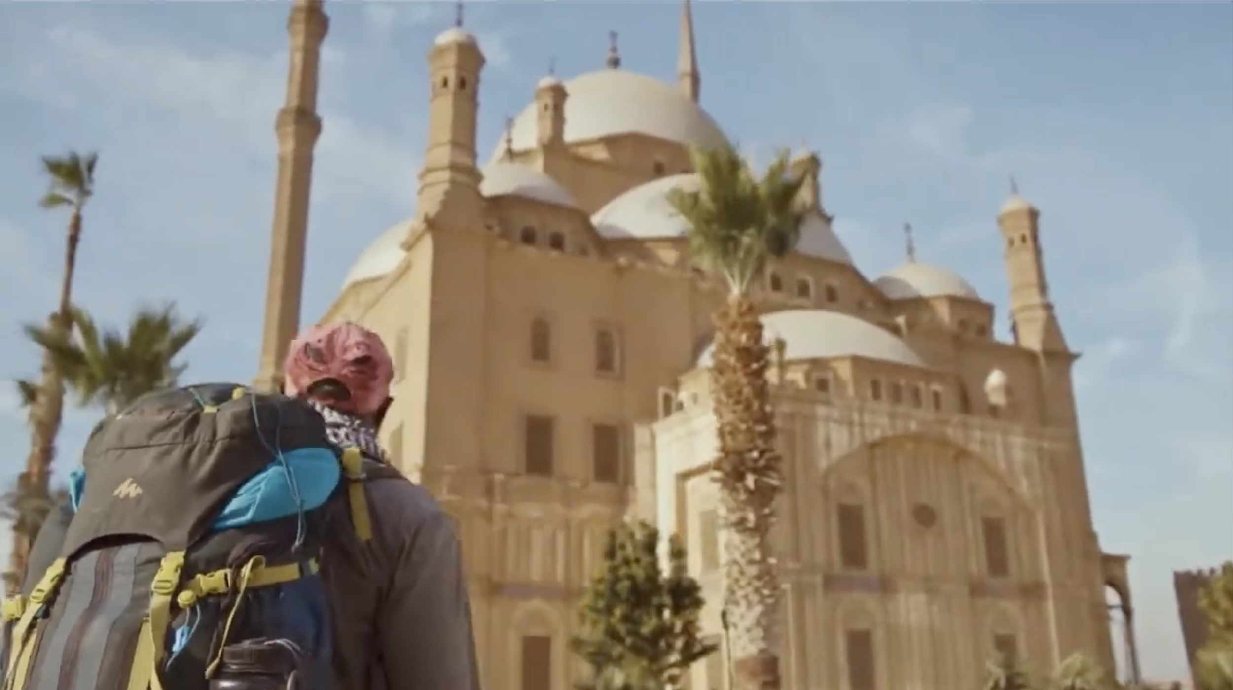 A person with pink hair, wearing a backpack, looks up at a large, historic, beige-colored castle with domes and towers, surrounded by palm trees, under a partly cloudy sky.
