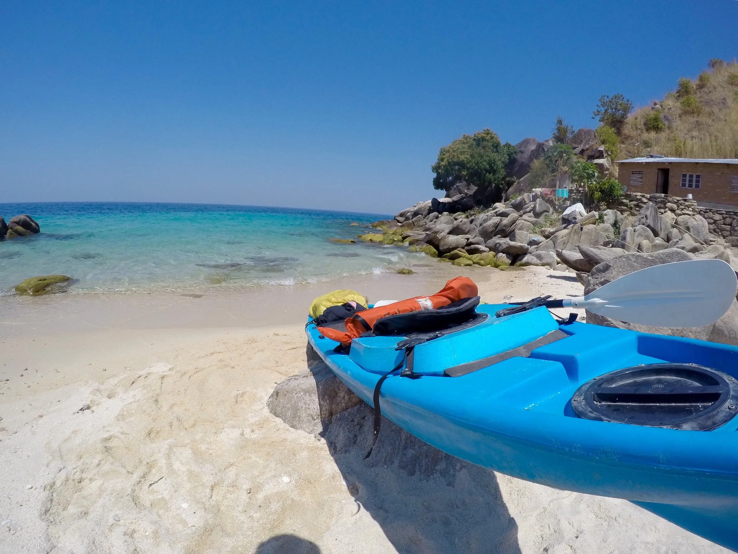 A kayak with paddles and gear resting on the sandy beach near the water's edge, with rocks, greenery, and a small house in the background under a clear blue sky.