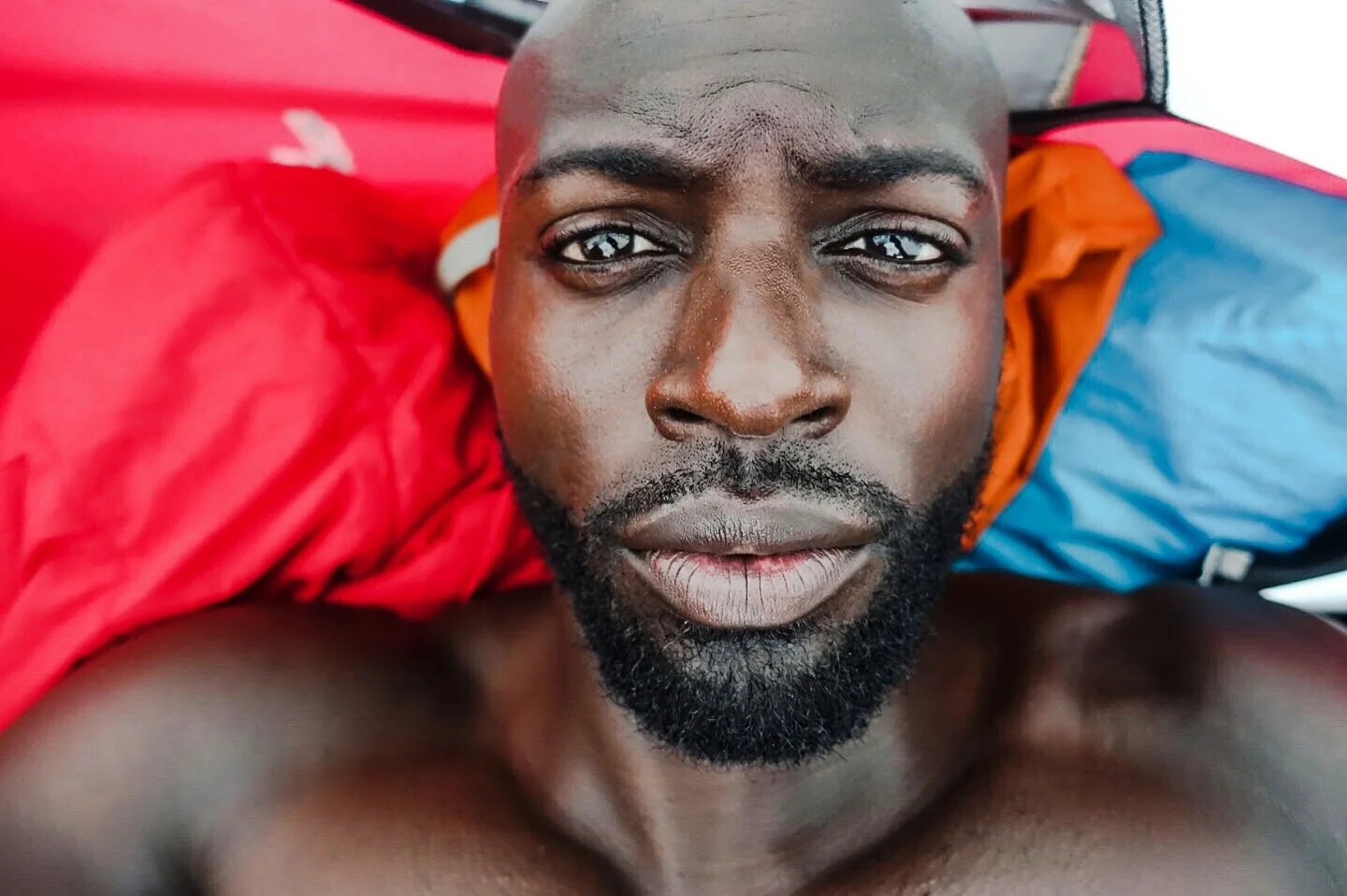Close-up of a shirtless man with a beard, lying on red and blue sleeping bags, looking into the camera.