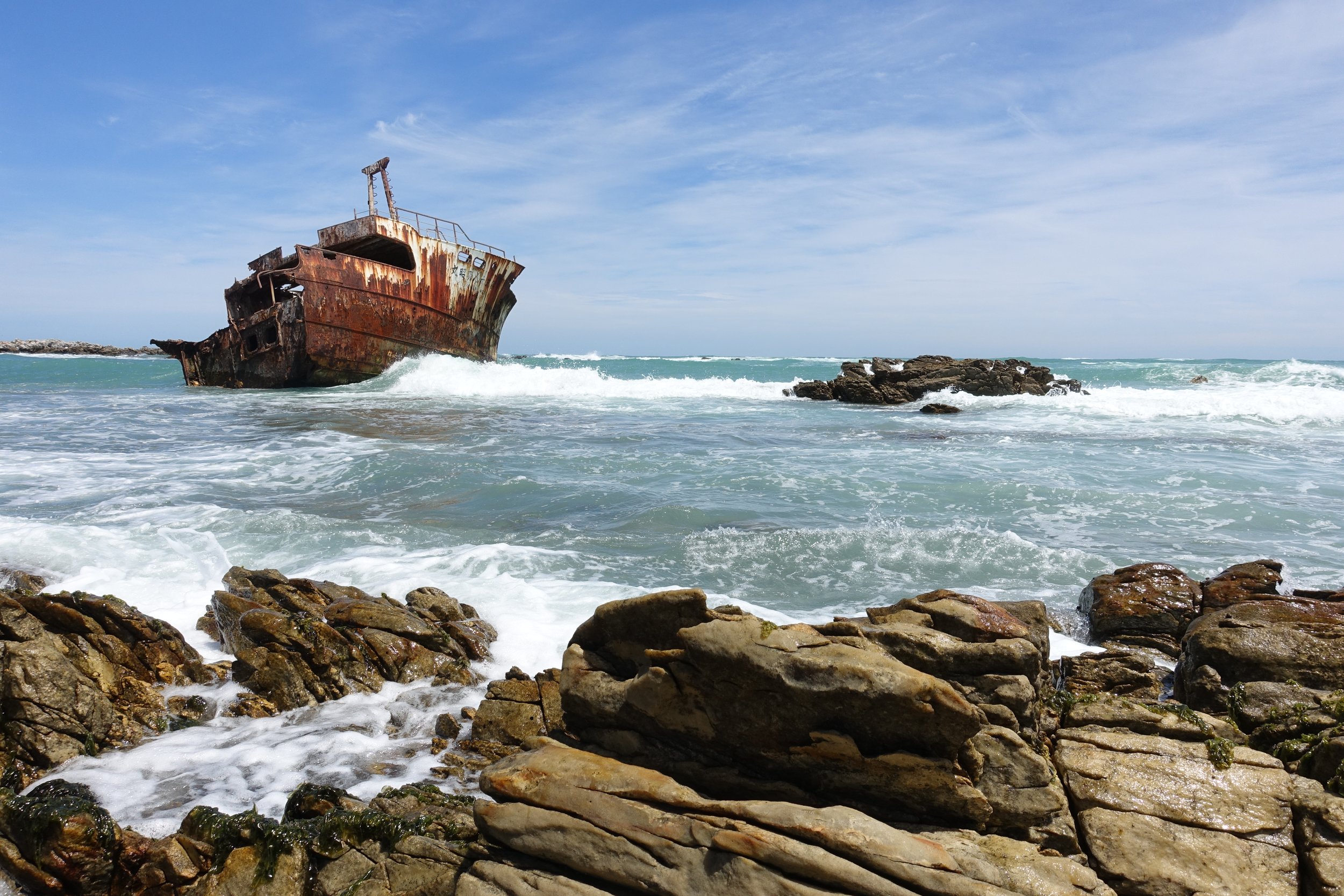 Rusty shipwreck on the rocky shoreline with blue sky and ocean waves.