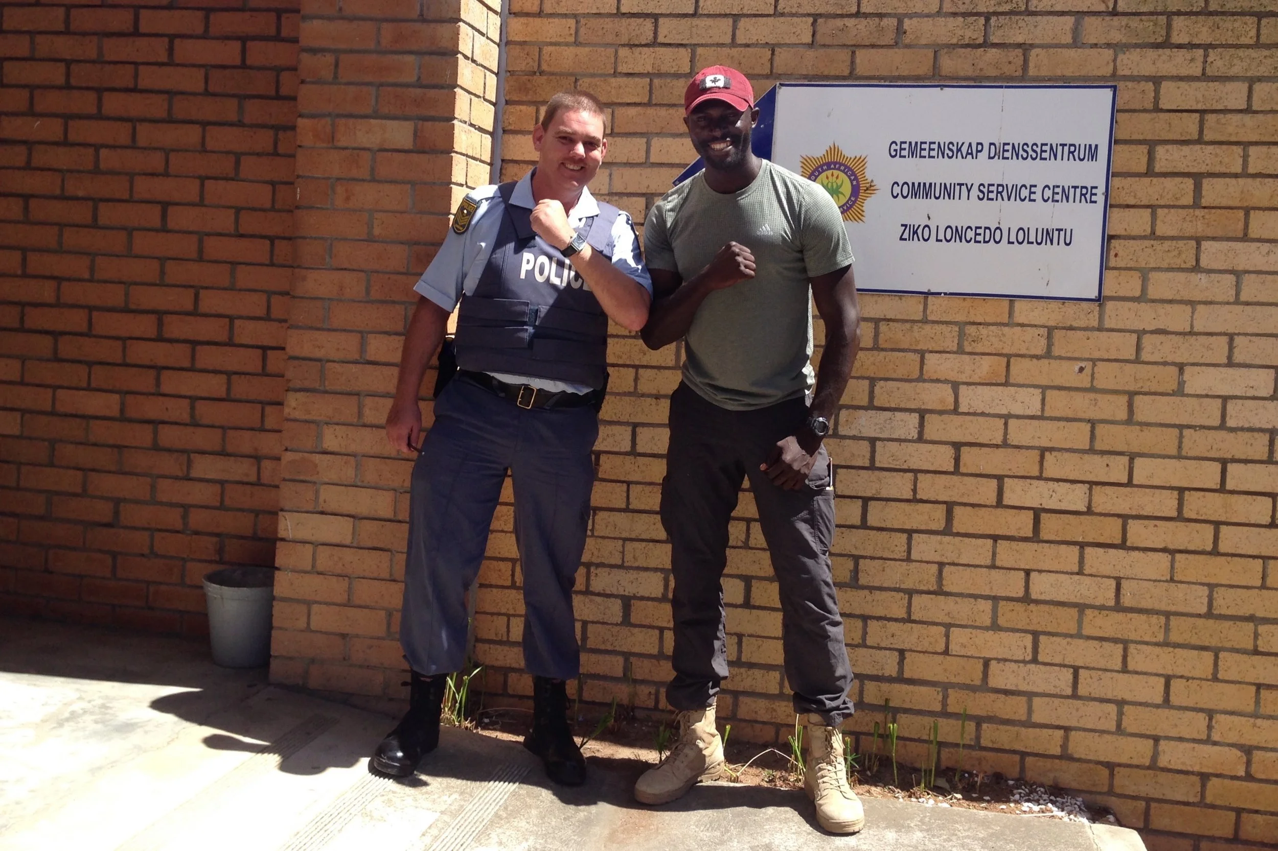 A police officer and a man standing outside a community service center, both smiling and making fists. The officer is in uniform, and the man is wearing casual clothing, including a red cap and beige boots.