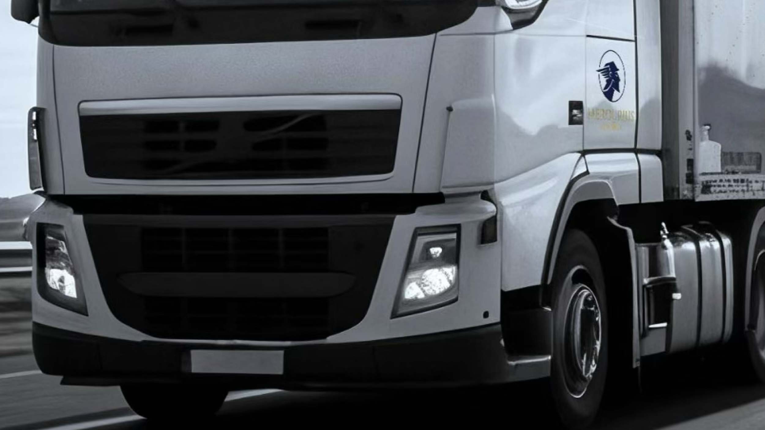 Camión de carga gris en movimiento, con logotipo en el costado y luces frontales encendidas.