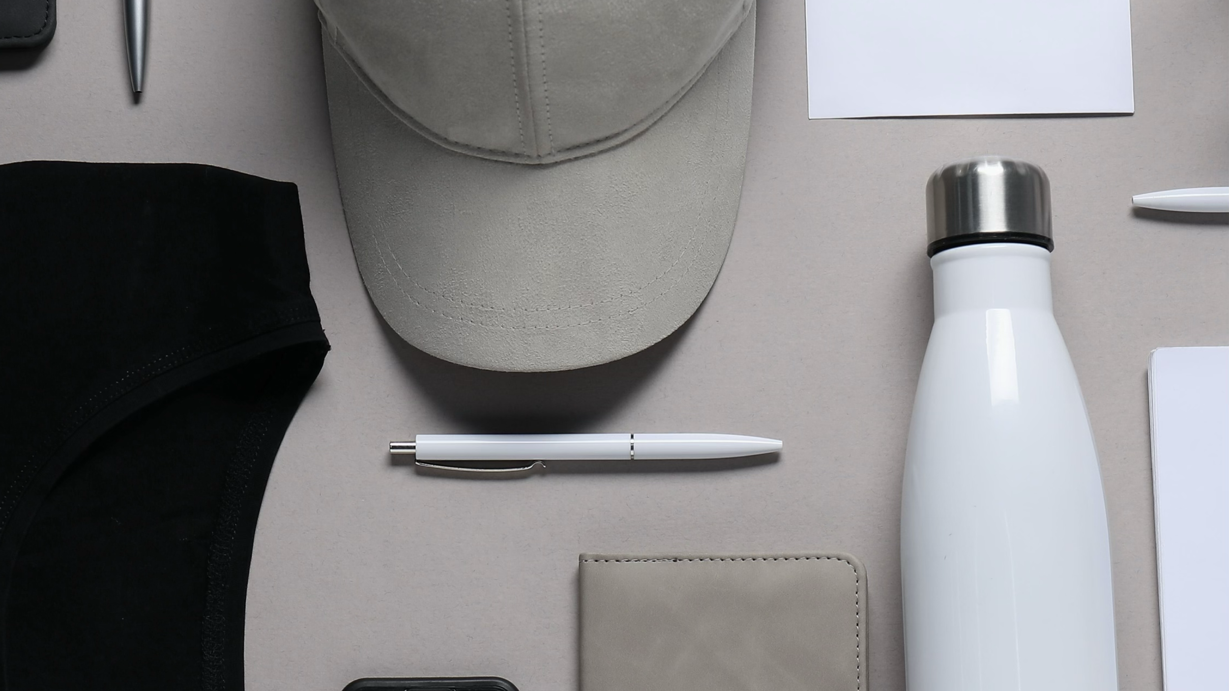 Vista superior de objetos de oficina y accesorios personales sobre una mesa, incluyendo una gorra gris, una botella blanca, un cuaderno beige, una pluma blanca, una computadora portátil negra, un recipiente termico, una hoja blanca, y un bolígrafo blanco.