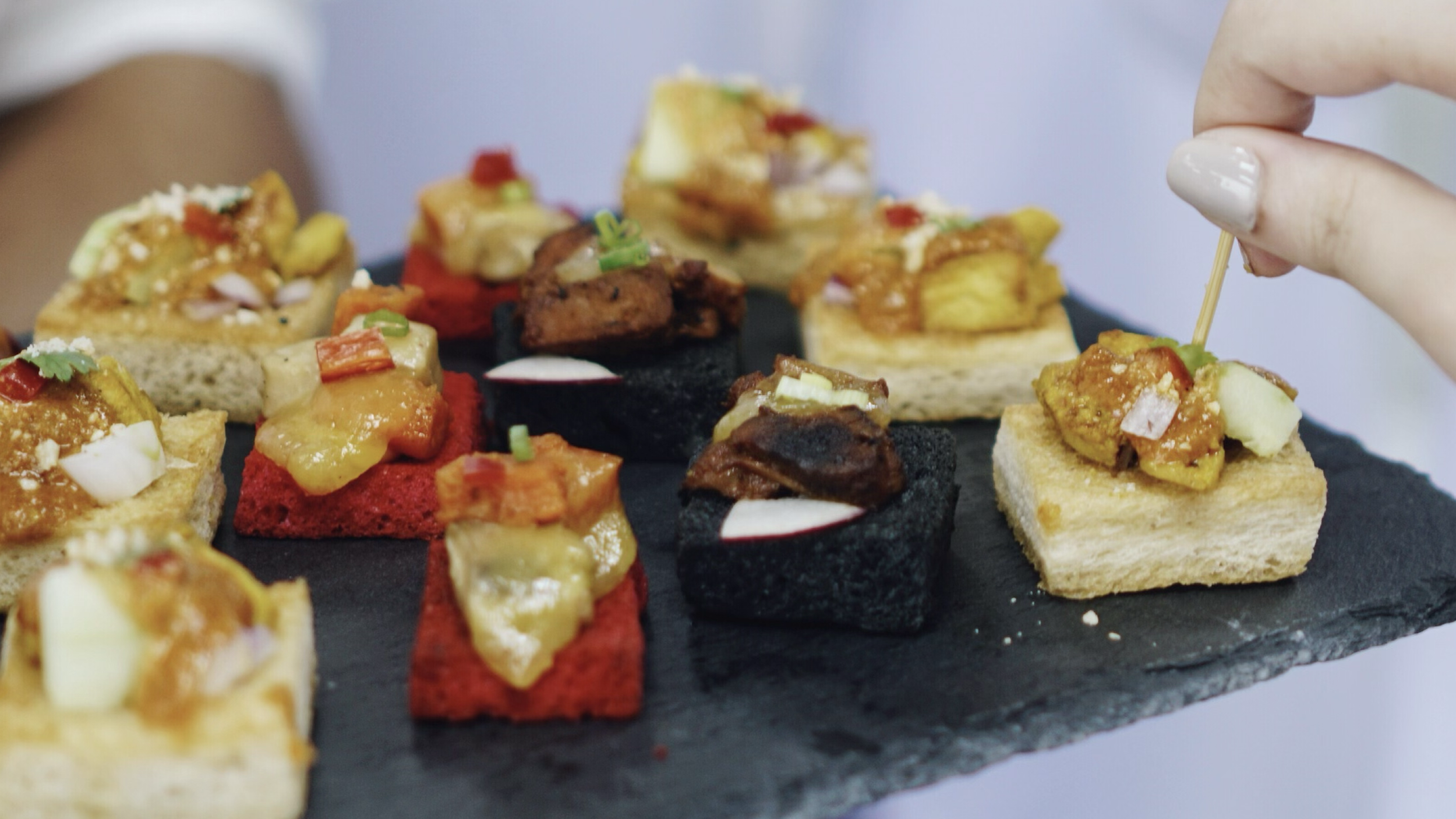 Variedad de canapés con diferentes ingredientes y colores servidos en una bandeja de piedra y una mano colocando uno con un palillo