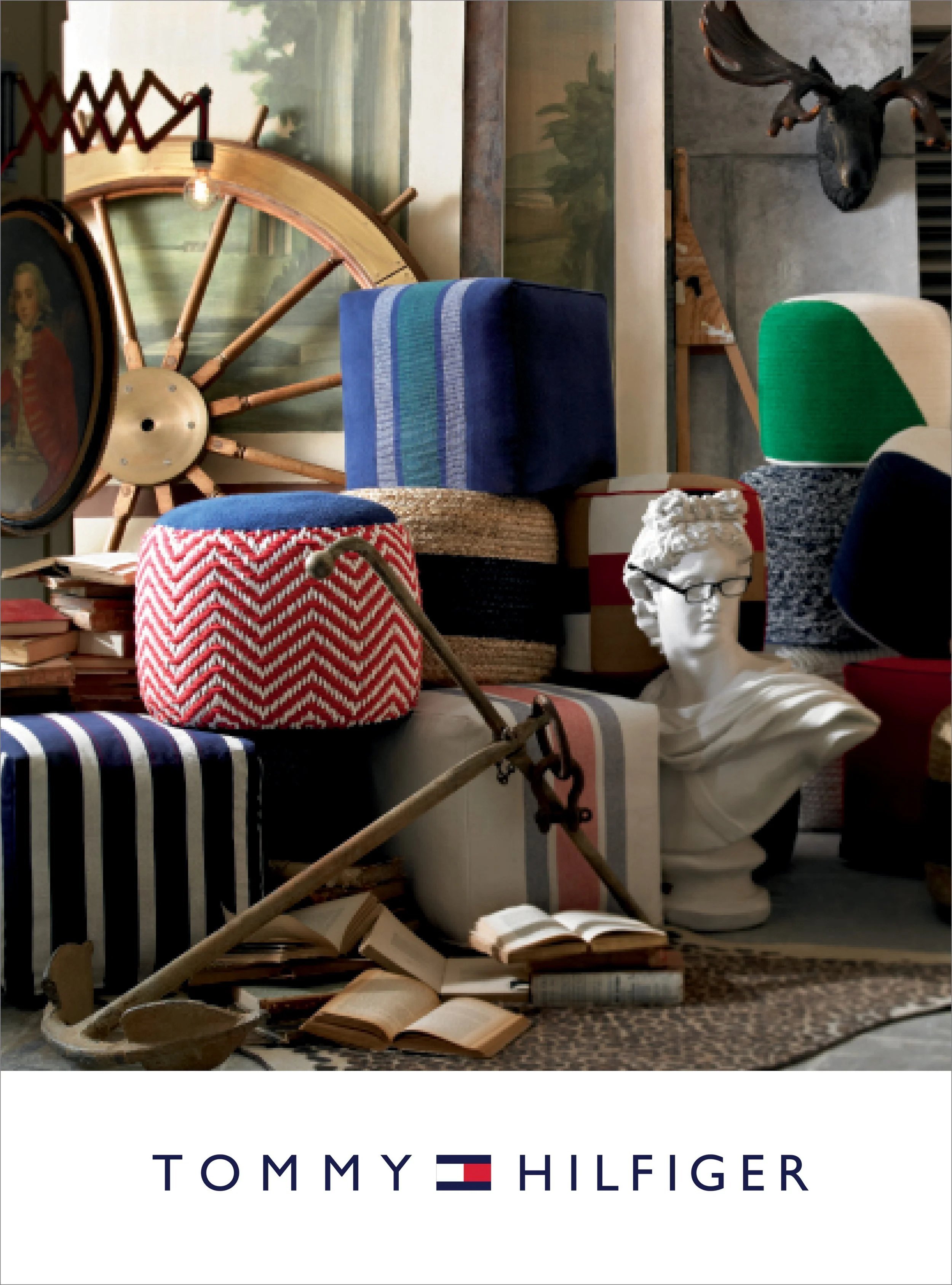 A collection of vintage and modern decor items including a wooden wheel, a blue and green upholstered chair, a bust sculpture, books, a painting of a landscape, a mounted deer head, and a sign with the Tommy Hilfiger logo.