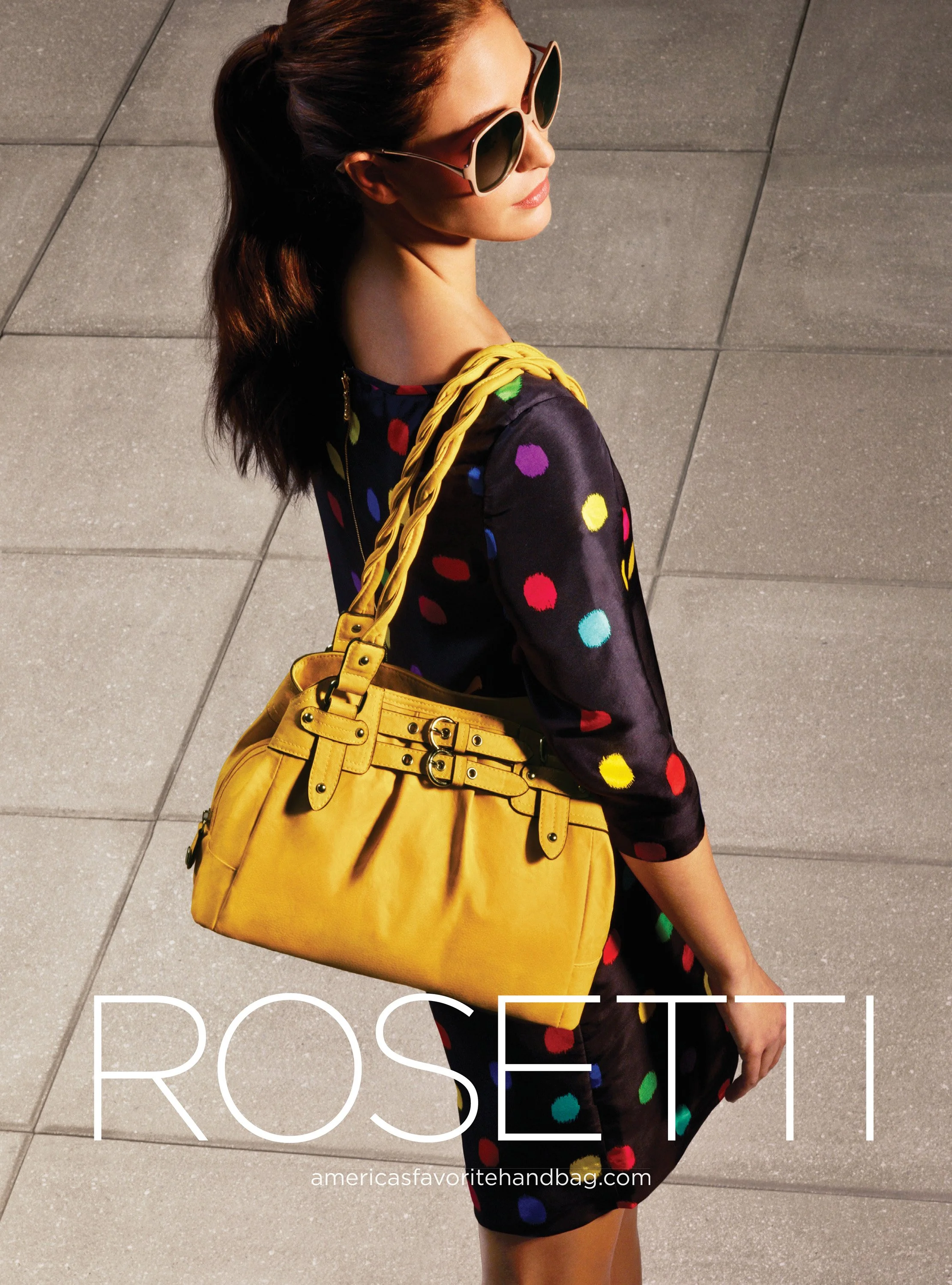 A woman with dark red hair wearing large sunglasses, a dark floral dress, and carrying a bright yellow handbag. She is captured from above against a tiled ground.