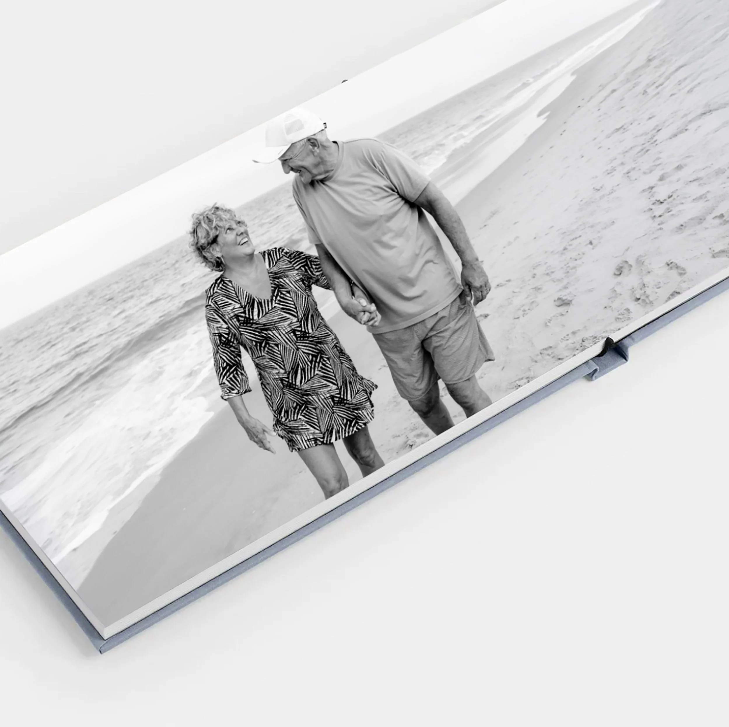 Black-and-white photograph of an elderly couple standing on the beach, holding hands, and smiling at each other with the ocean in the background.