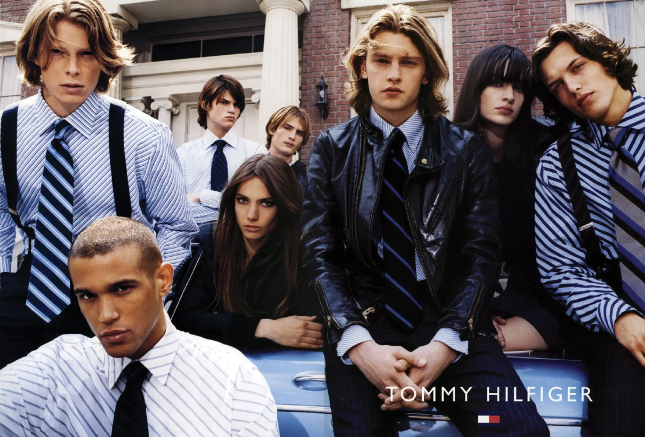 Group of young men and women, dressed in stylish clothing, standing in front of a brick building with columns. They have serious expressions, with some looking directly at the camera. The image features the Tommy Hilfiger logo at the bottom.