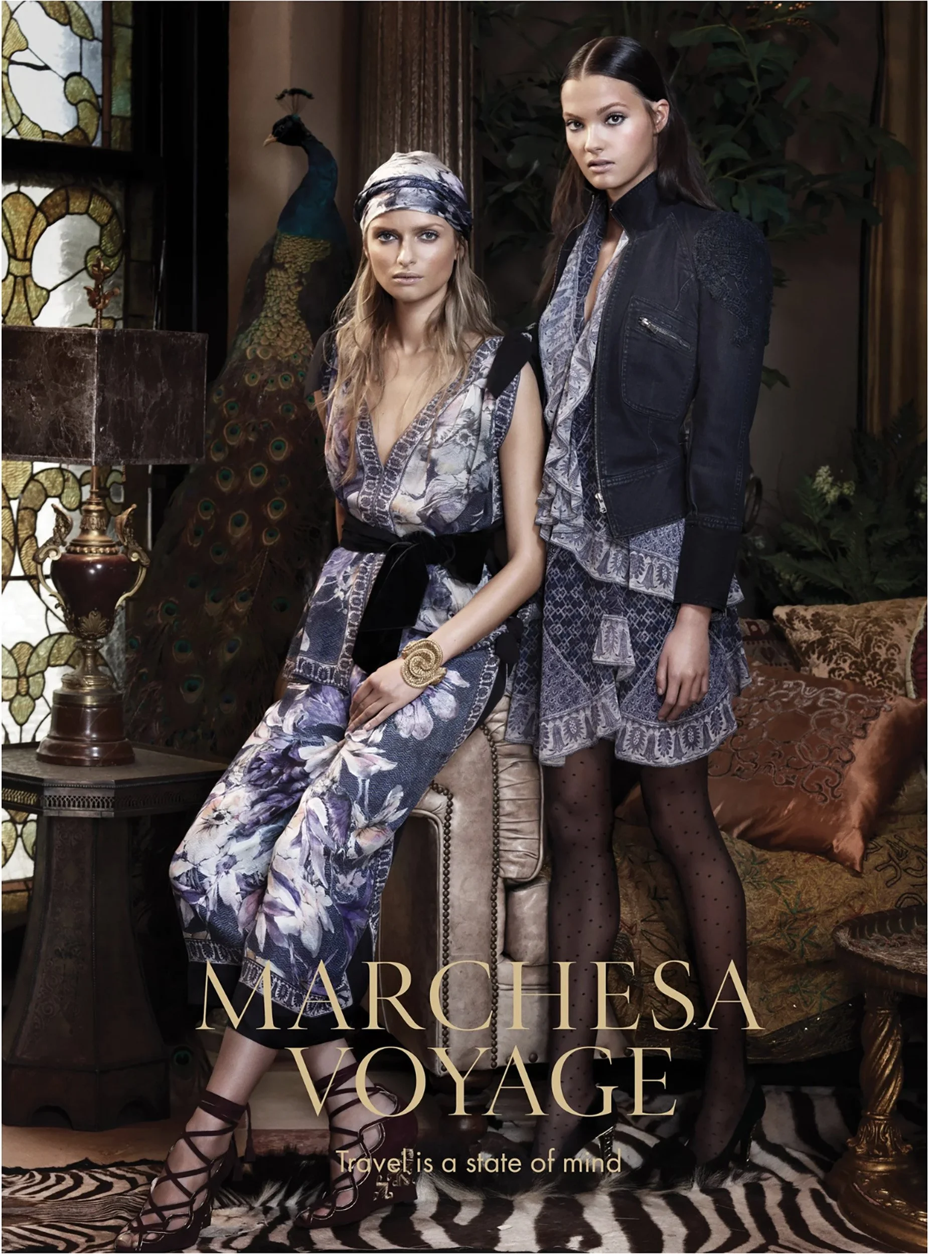 Two women inside a decoratively furnished room, standing near a vintage-style table with a peacock statue in the background. One woman is seated wearing floral pants, a floral top, and a wrap-around headscarf. The other woman stands wearing a black j