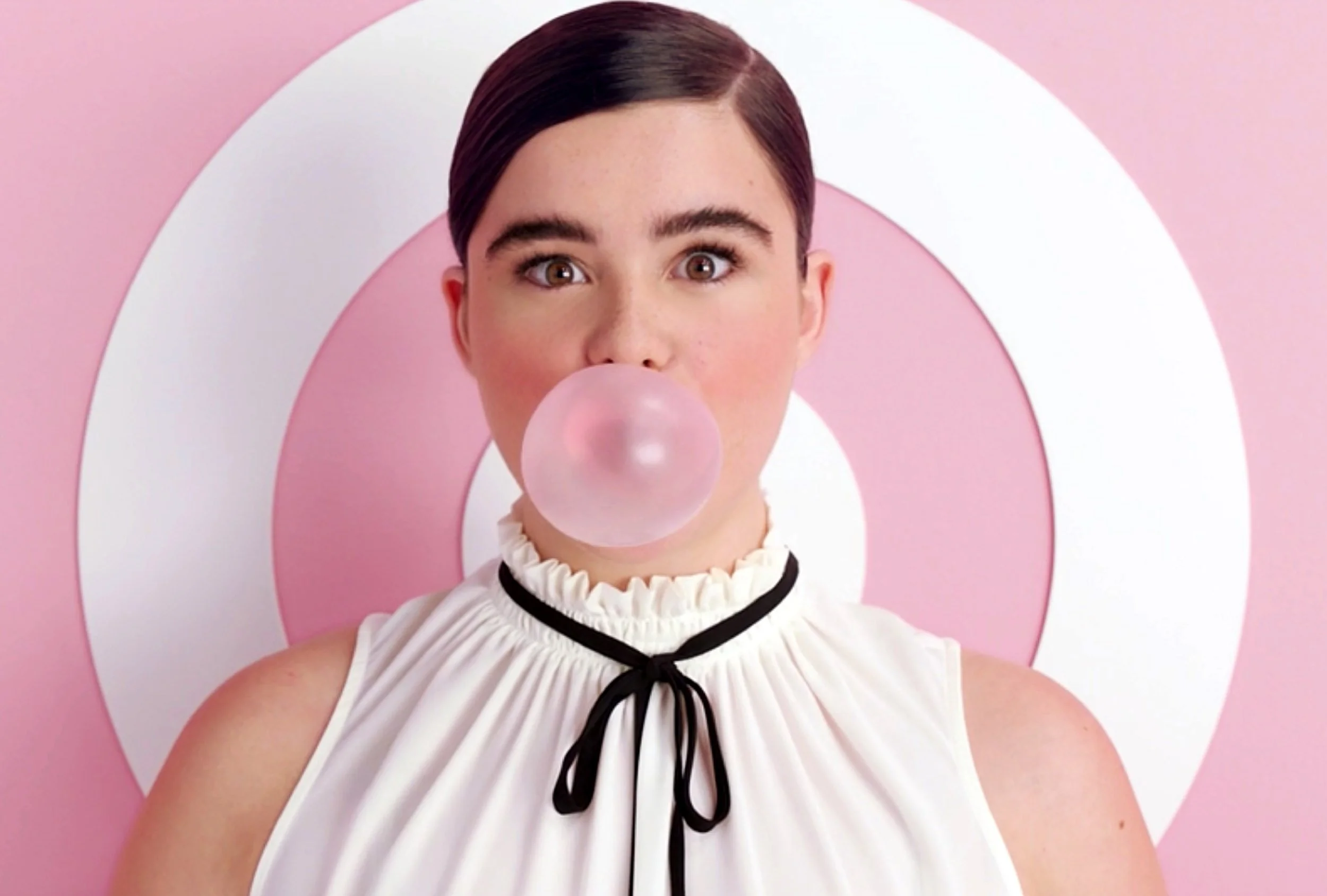 A woman with short dark hair wearing a white dress with ruffled neckline and black ribbon, blowing bubblegum in front of a pink and white target background.