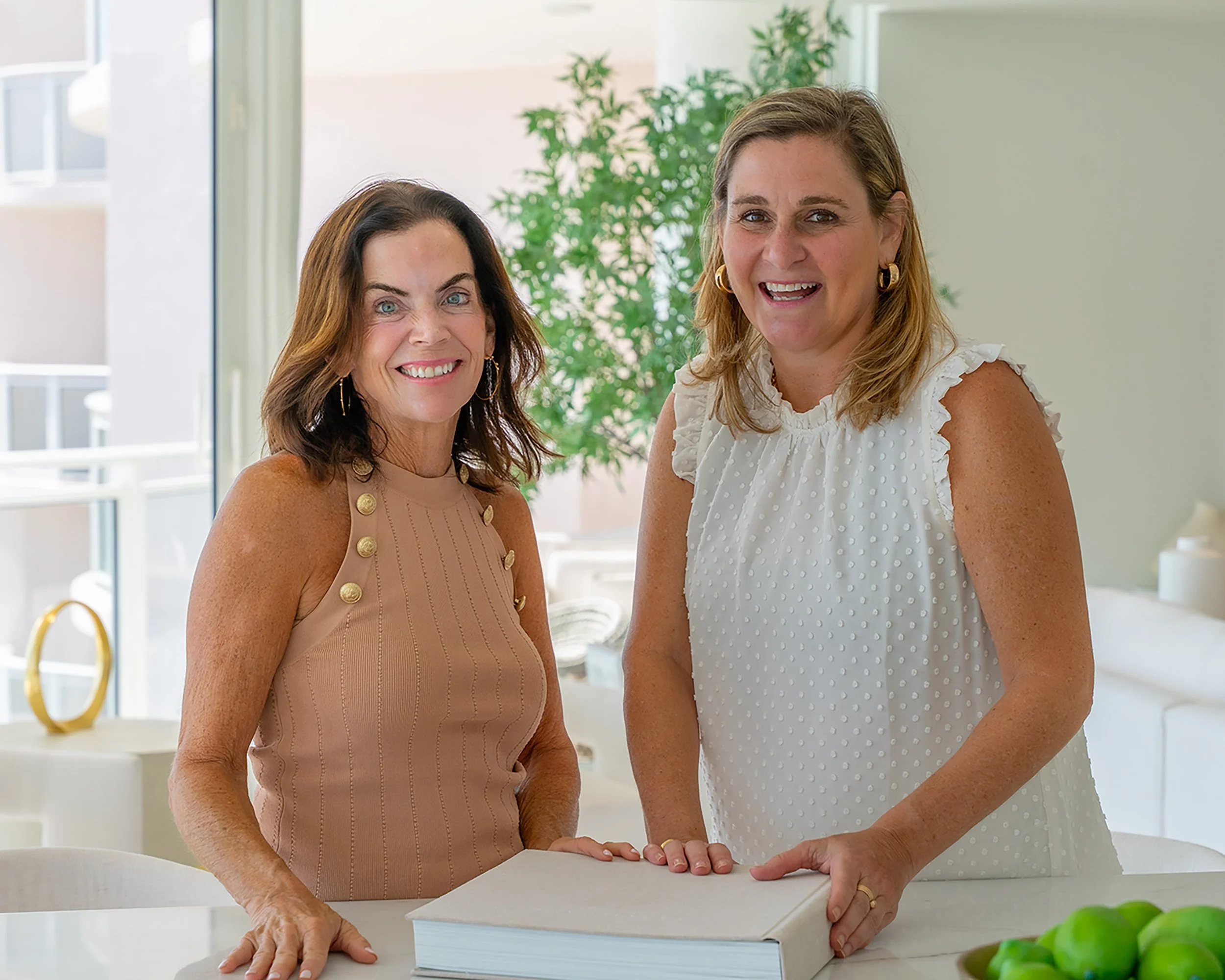 Two women smiling and standing together in a bright room with large windows and green plants, one is wearing a sleeveless beige top with gold button details, and the other is wearing a white sleeveless top with ruffled shoulders.