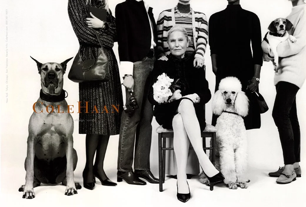Black and white photo of six women and two dogs, with one woman sitting holding a bouquet of flowers. The women are dressed in stylish clothing, and the dogs are sitting or being held.