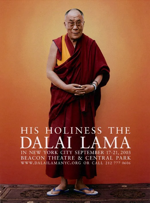 A monk dressed in red robes standing on a carpet, smiling with his hands clasped in front of him, against an orange background with promotional text about a Dalai Lama event in New York City in 2003.
