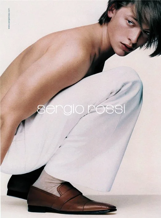 A shirtless young man with dark hair crouching in a studio with a plain, light background. Text reads "sergio possi".